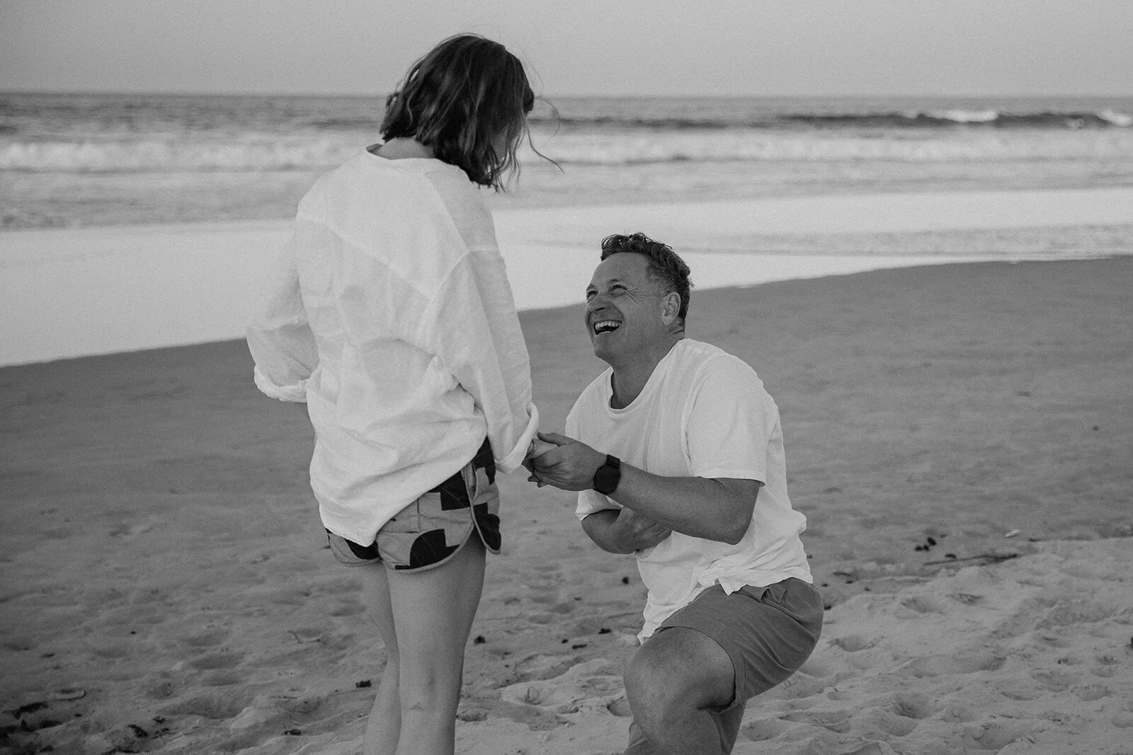 Black and white of the man laughing on one knee with the ring, Jamie and Jessica's beach marriage proposal at Belongil Beach, Byron Bay