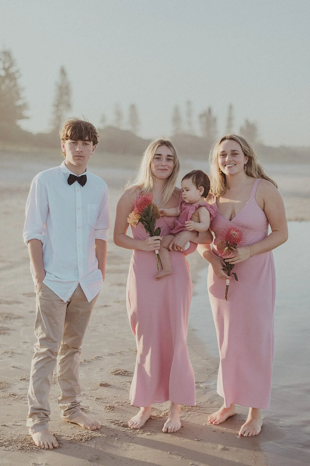 Teenage boy and two young women in pink dresses standing on the sand, Maritess and Mark's elopement at Lennox Head