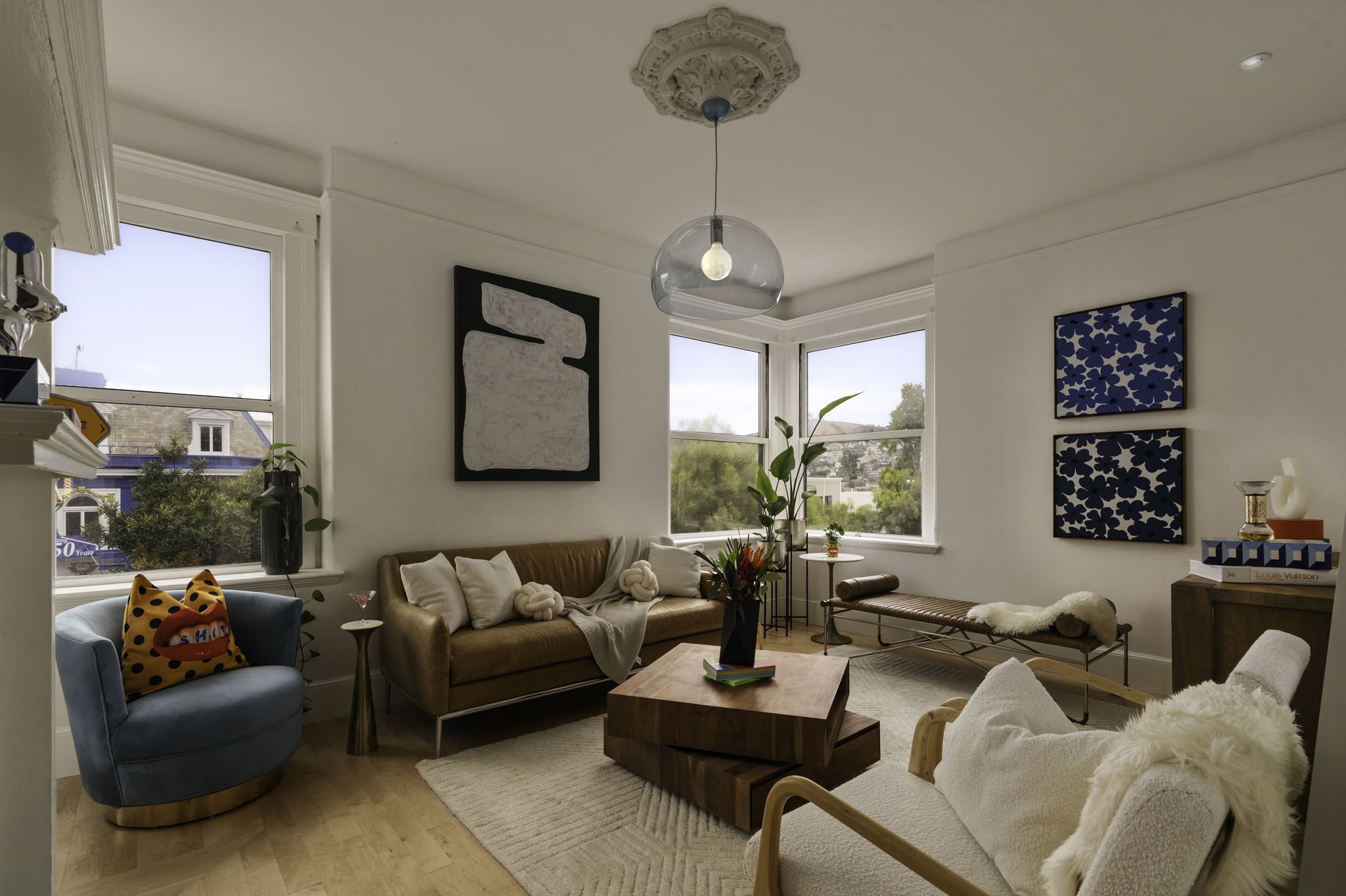 A cozy living room with beige and blue furniture, including a beige sofa with pillows, a blue armchair with a decorative pillow, and a wooden coffee table. The room features large windows letting in natural light, with artwork hanging on the walls, and potted plants for decor.