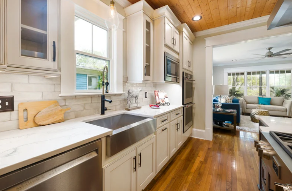 Bright kitchen with white cabinets, marble countertops, black faucet, and stainless steel sink, leading to a living room with a blue sofa and large windows.