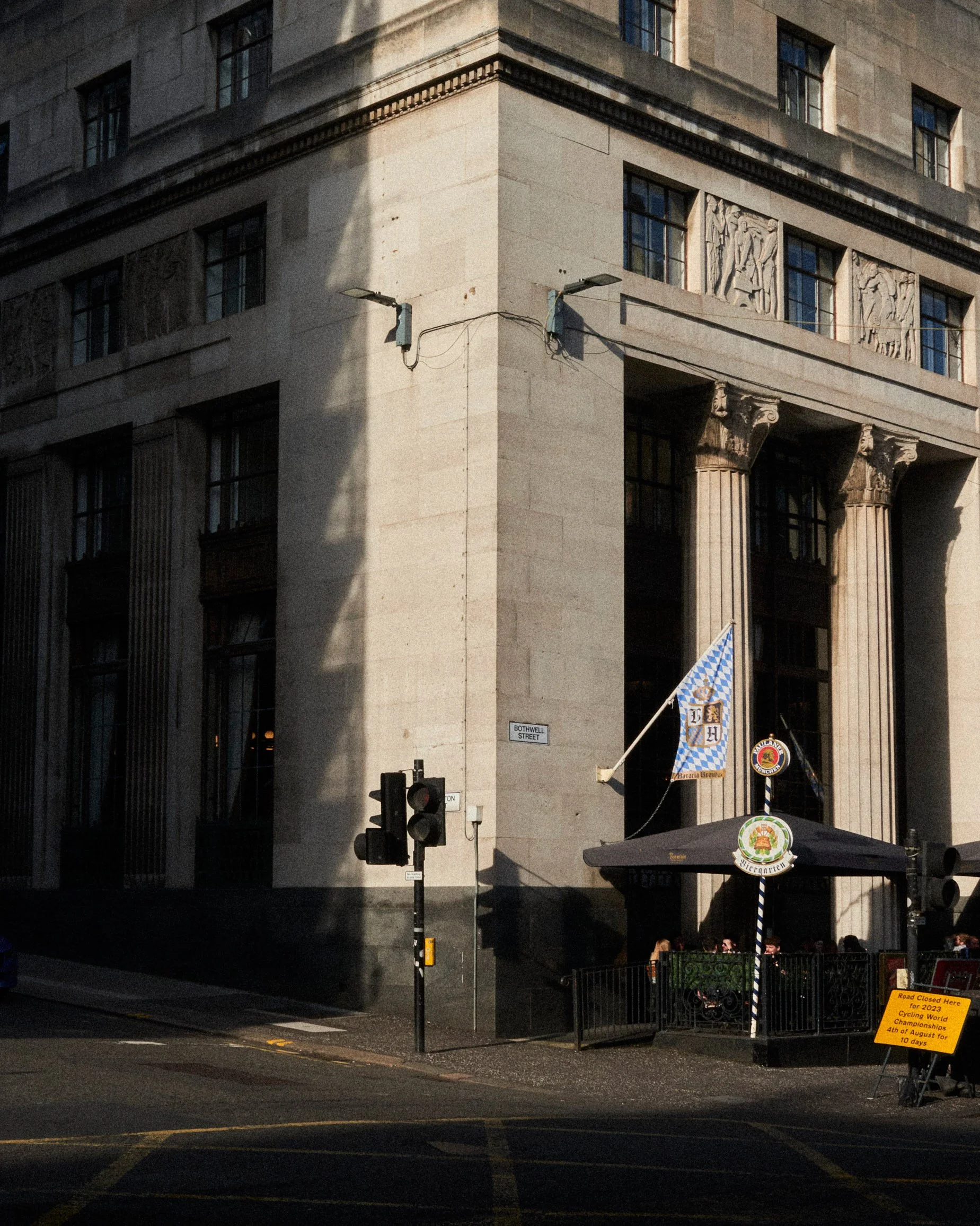 Photo d’un bâtiment historique avec des colonnesion et des sculptures au niveau du toit, angle de la rue avec des lampadaires et une bannière aux couleurs bleu et blanc, avec un drapeau bavarois, indication d’un événement sportif à venir en août