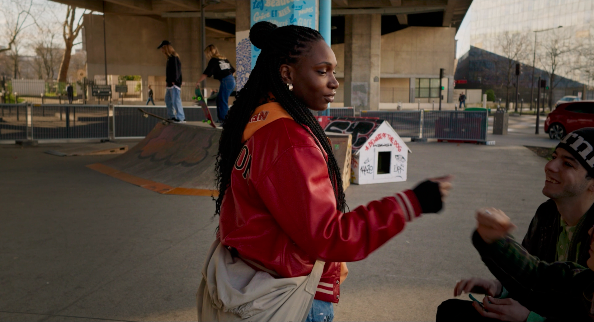 Une femme noire avec de longues tresses, portant une veste rouge et un sac beige, échange un sourire avec un homme portant un bonnet noir et un sweat noir, dans un skatepark avec enfants en arrière-plan.