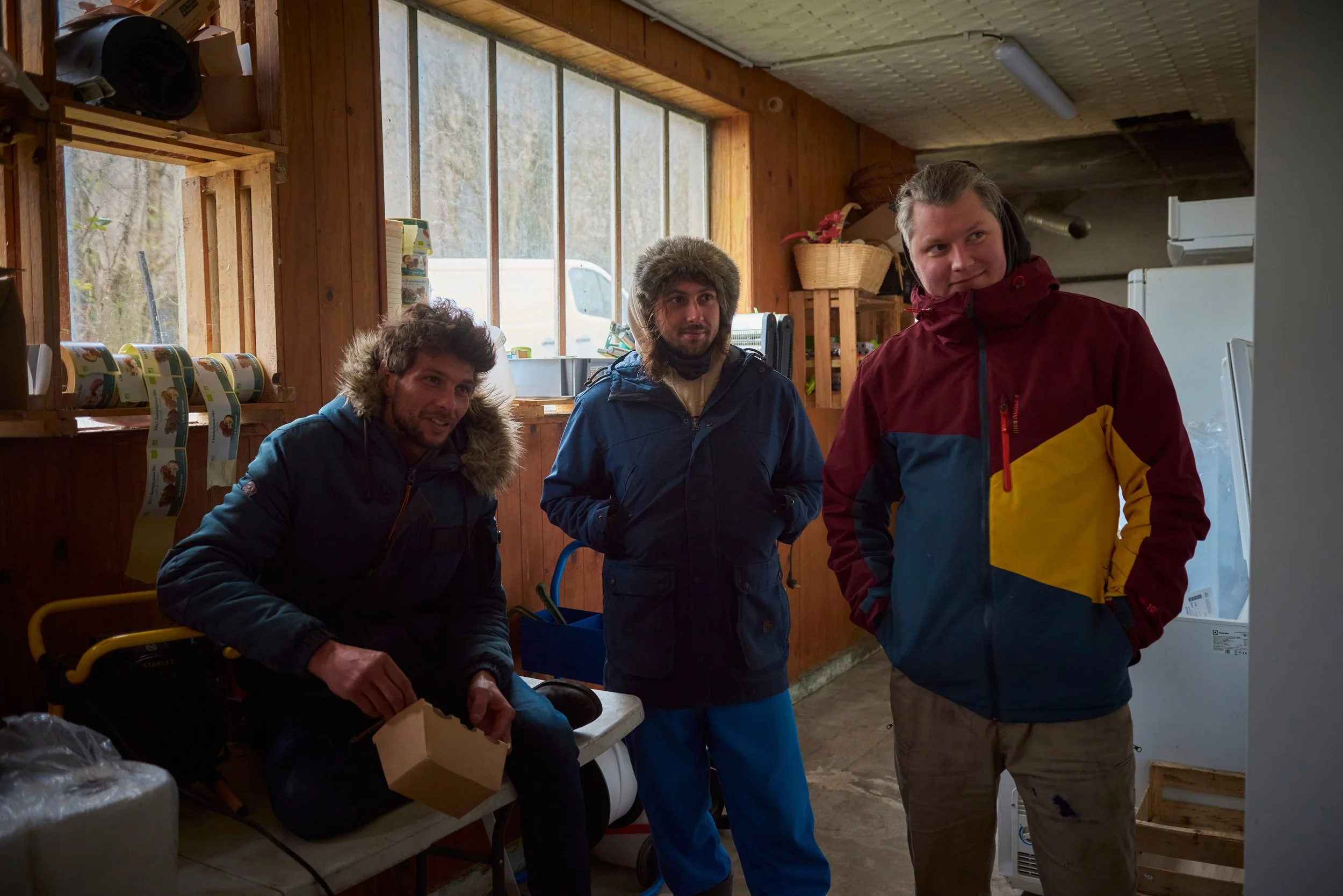 Trois hommes portent des vêtements d'hiver dans une pièce en bois avec de grandes fenêtres. L'un d'eux est assis, les autres se tiennent debout.