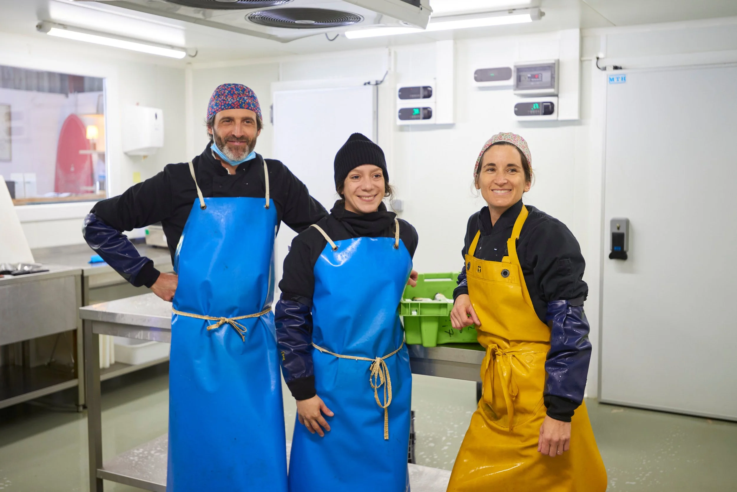 Trois personnes vêtues de vêtements de protection en combinés bleus ou jaunes dans une cuisine ou une usine alimentaire, avec des équipements en arrière-plan.