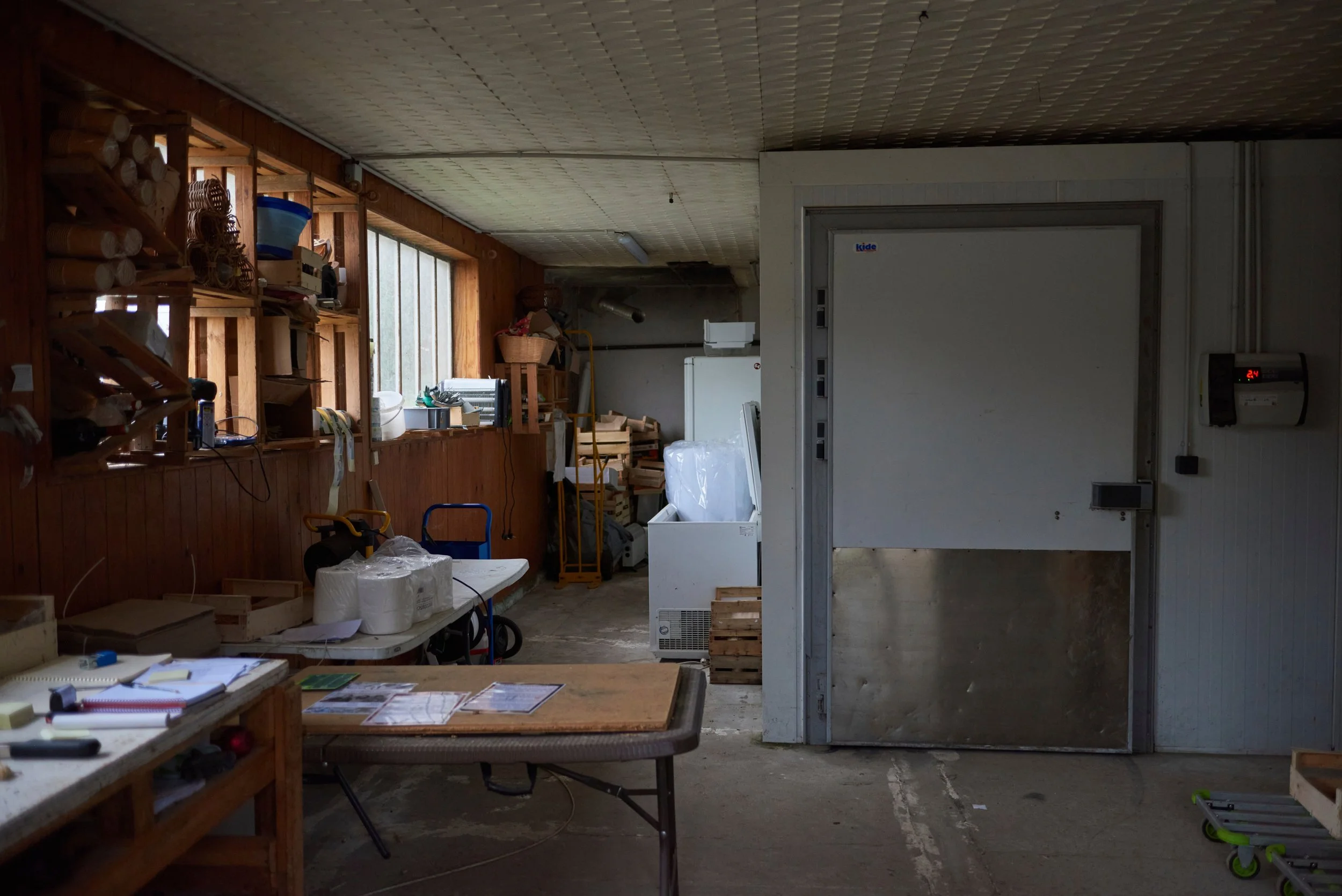 Un atelier ou un garage avec des étagères en bois remplies d'outils, de matériaux et de divers objets, une table de travail avec des papiers et des outils, et un espace vide avec un mur blanc et un porte frigorifique.