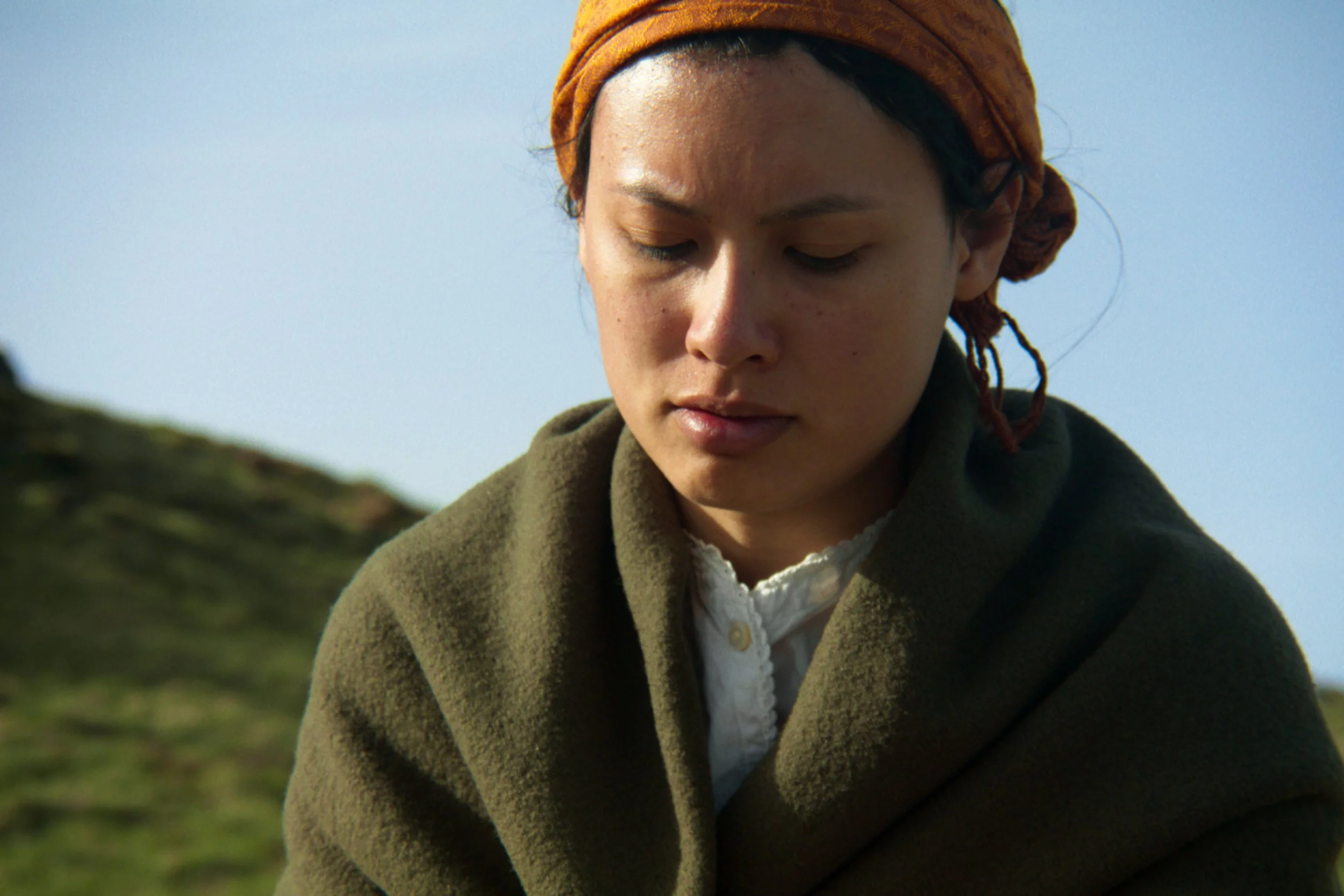 Une femme avec un turban orange, portant un manteau vert foncé et une chemise blanche, regardant vers le bas dans un paysage en plein air