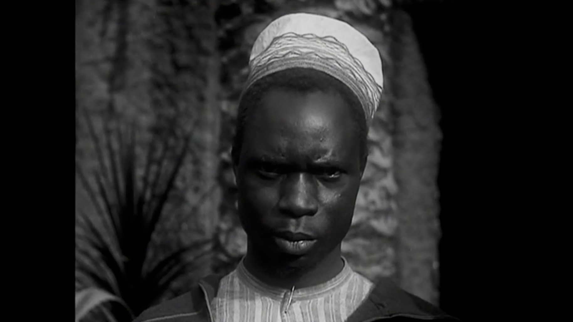 Portrait noir et blanc d'une jeune femme africaine portant un foulard blanc sur la tête, avec un fond flou de nature ou de pierre.