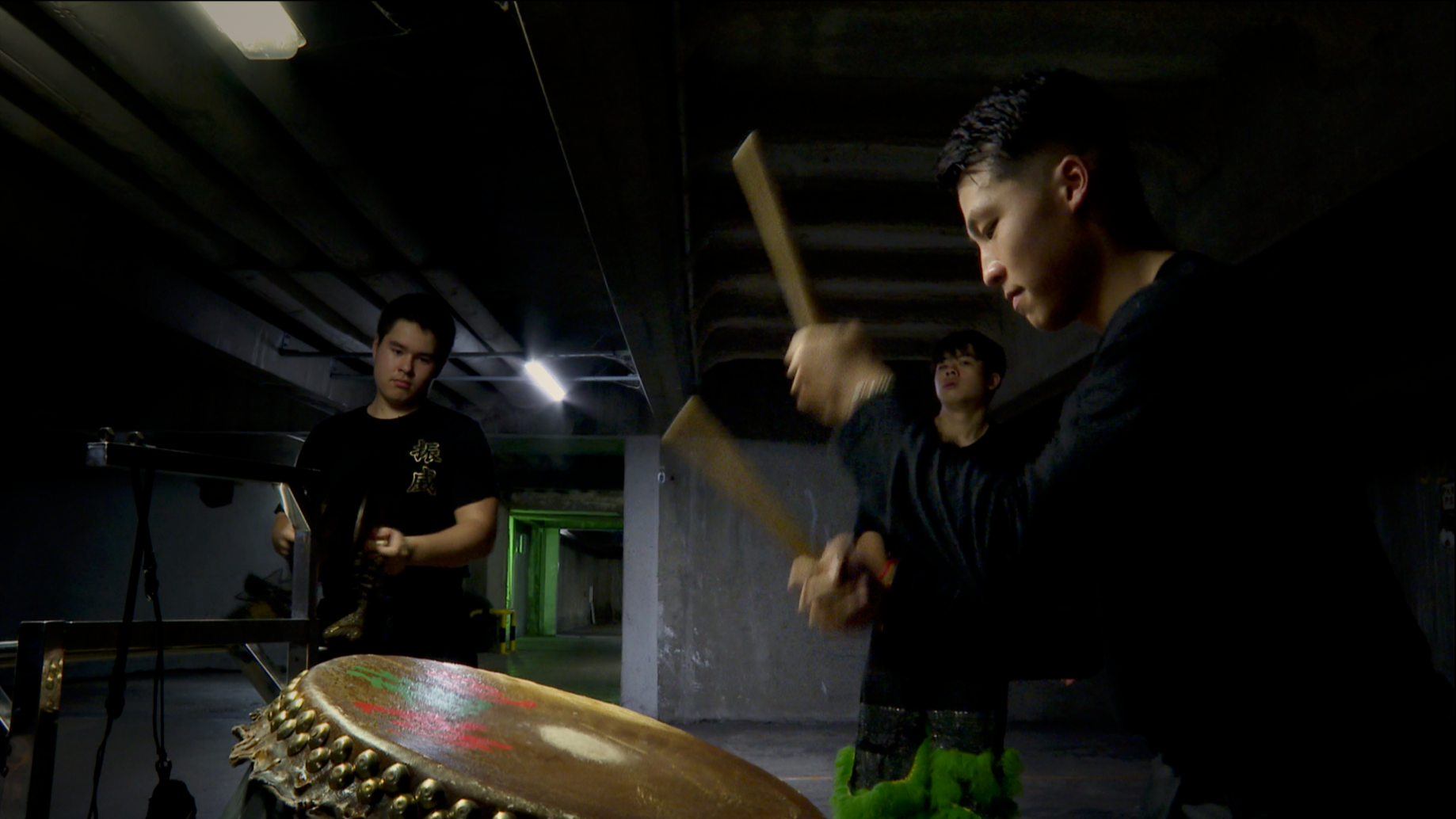 Trois jeunes hommes jouent de la musique dans un endroit sombre, une personne frappe un tambour et deux autres tiennent des instruments, un guzheng et un instrument à cordes.