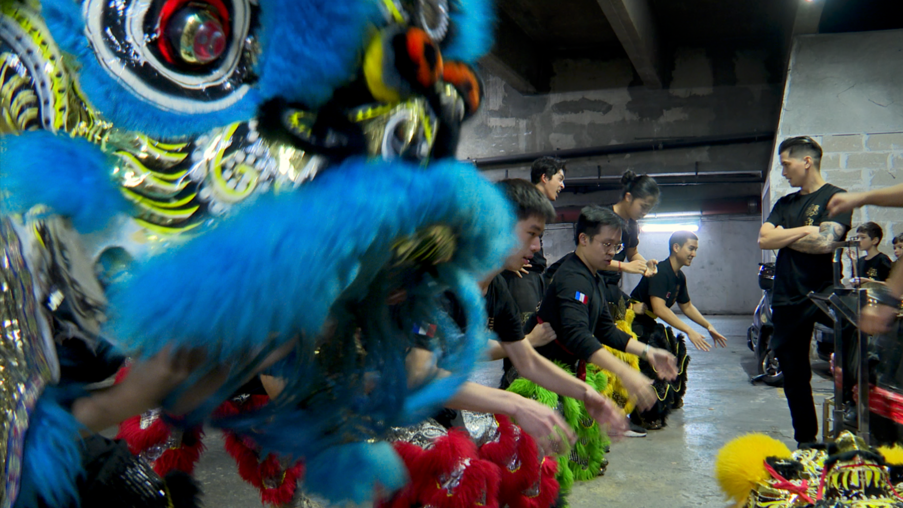 Groupe de personnes assises au sol en costume traditionnel, préparant un spectacle de danse, avec un lion de costume bleu dans le premier plan.