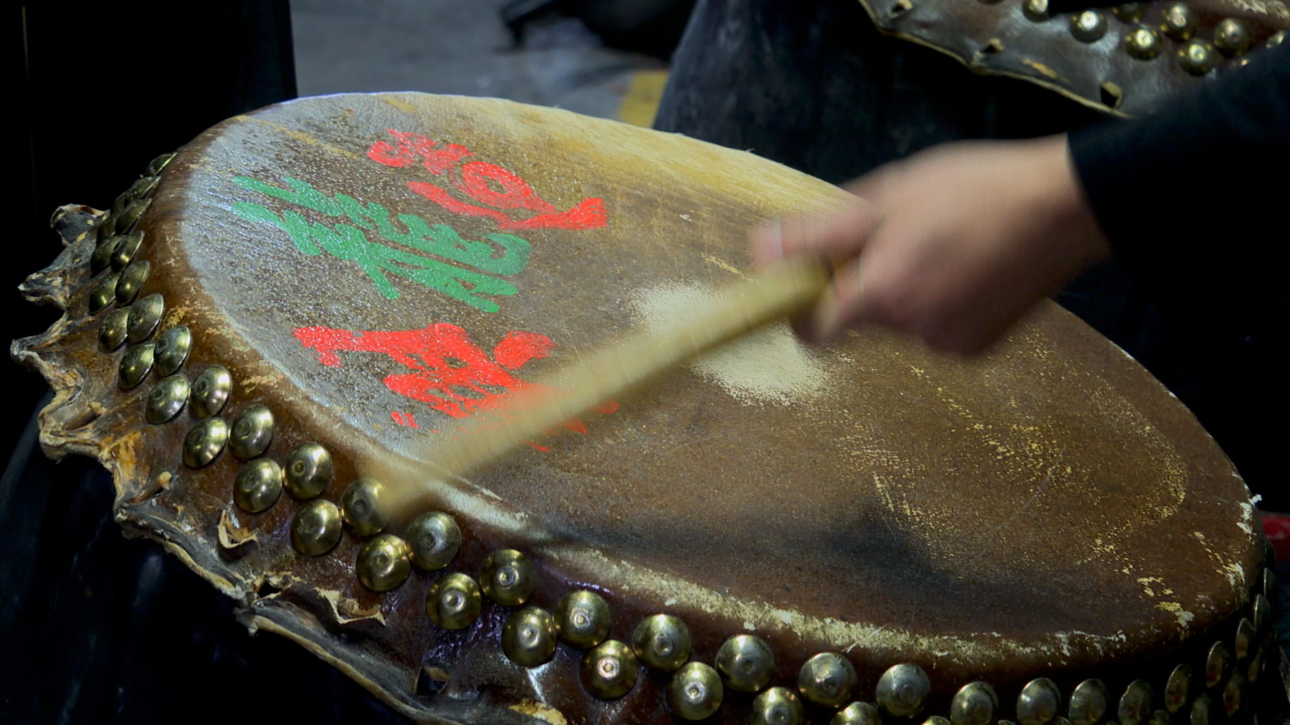 Une main joue du tambour avec un bâton sur un tambour traditionnel orné de clous en métal, avec des motifs rouges et verts peint sur la surface.