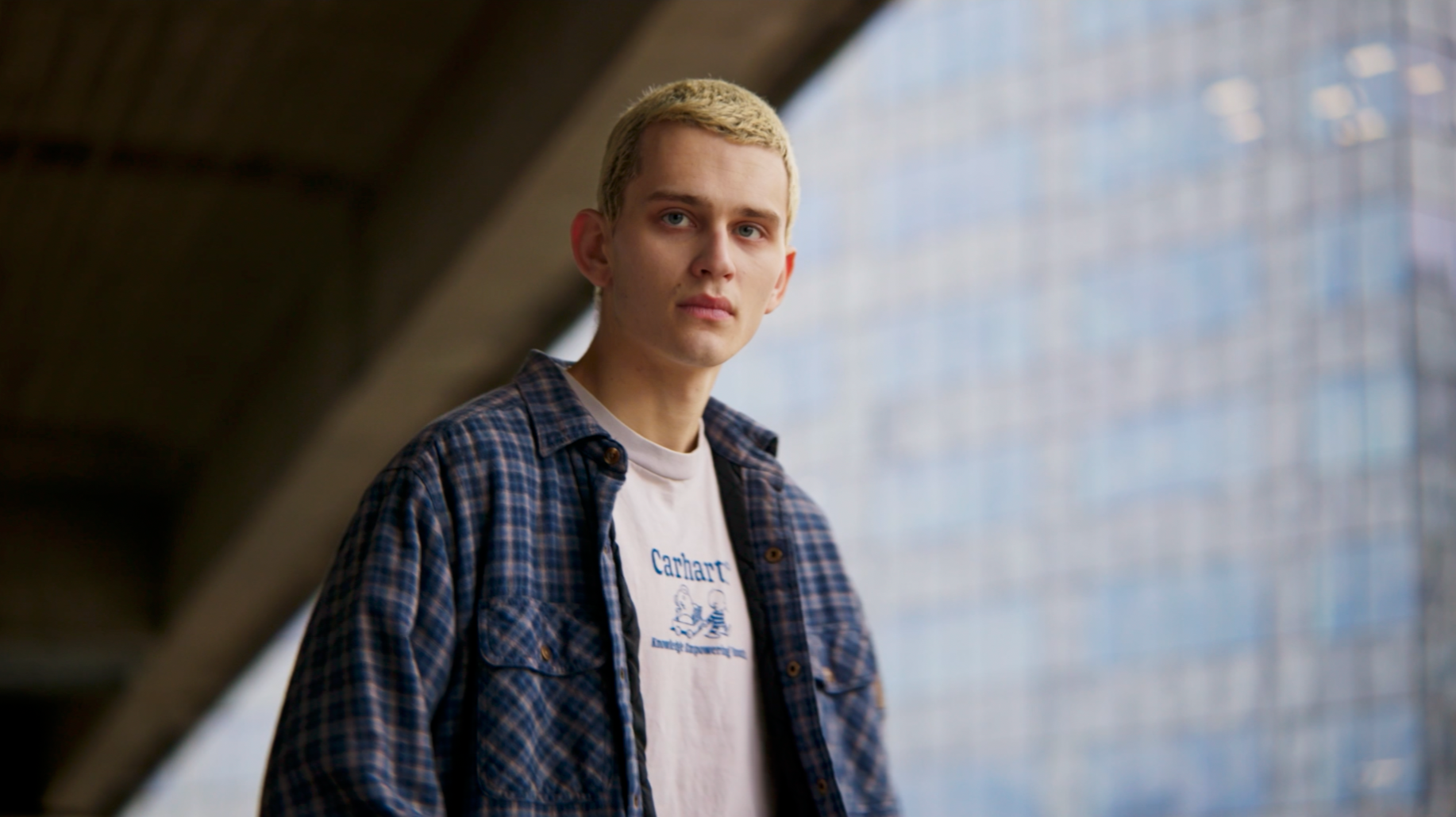 Jeune homme avec des cheveux blonds, portant une chemise à carreaux et un t-shirt "Carhartt", regardant vers la caméra dans un environnement urbain.