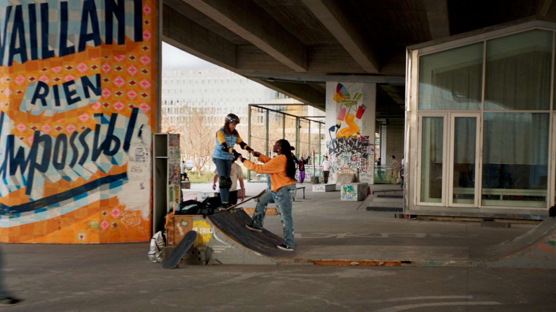 Deux jeunes femmes, une portant un casque et l'autre en orange, s'entraînent au skateboard dans un skatepark sous un pont couvert, avec des graffitis sur les murs.