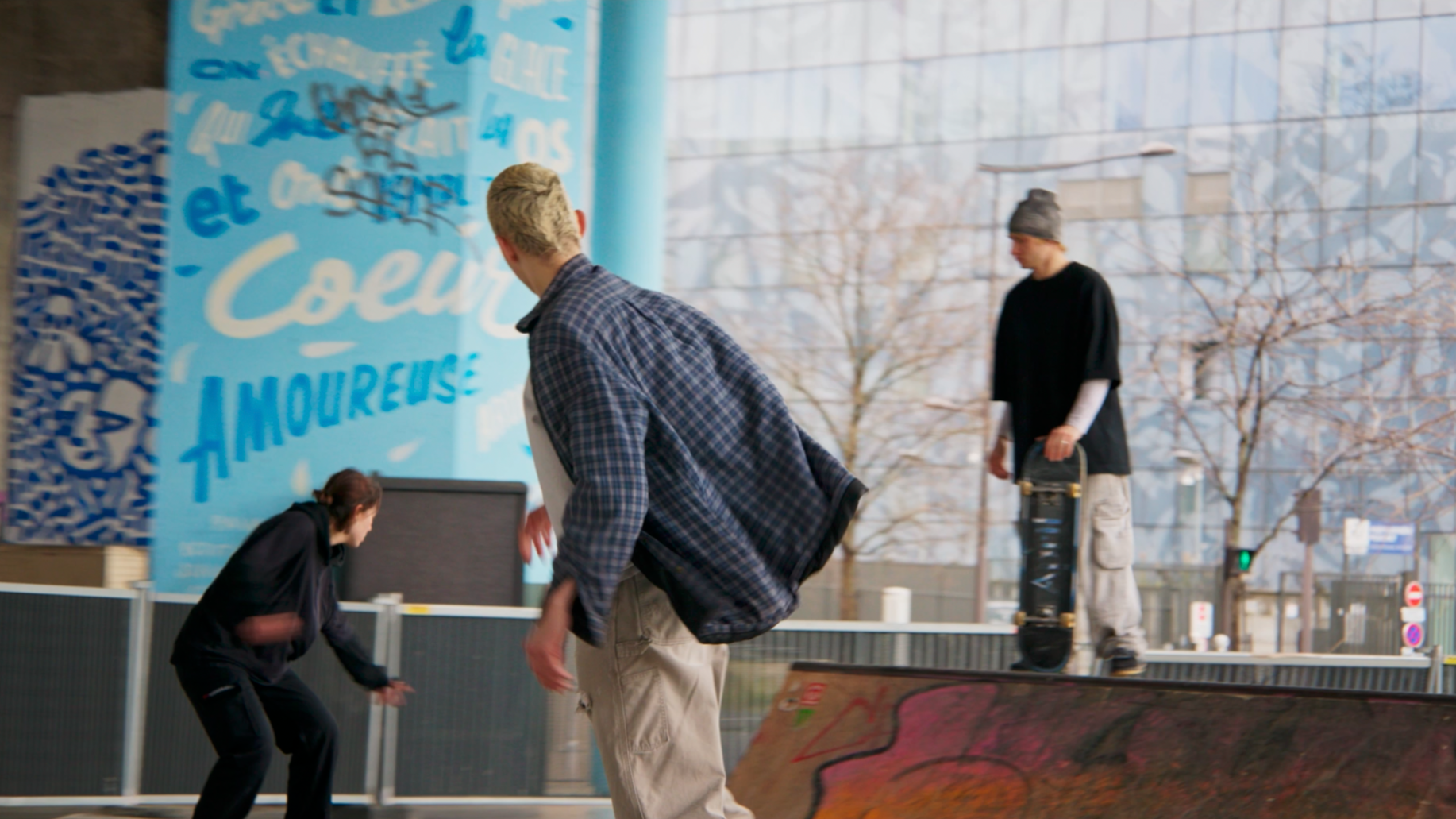 Trois jeunes gens dans un skatepark urbain. Deux d'entre eux sont debout et une est en position de squat ou de chute, tous portant des vêtements décontractés.