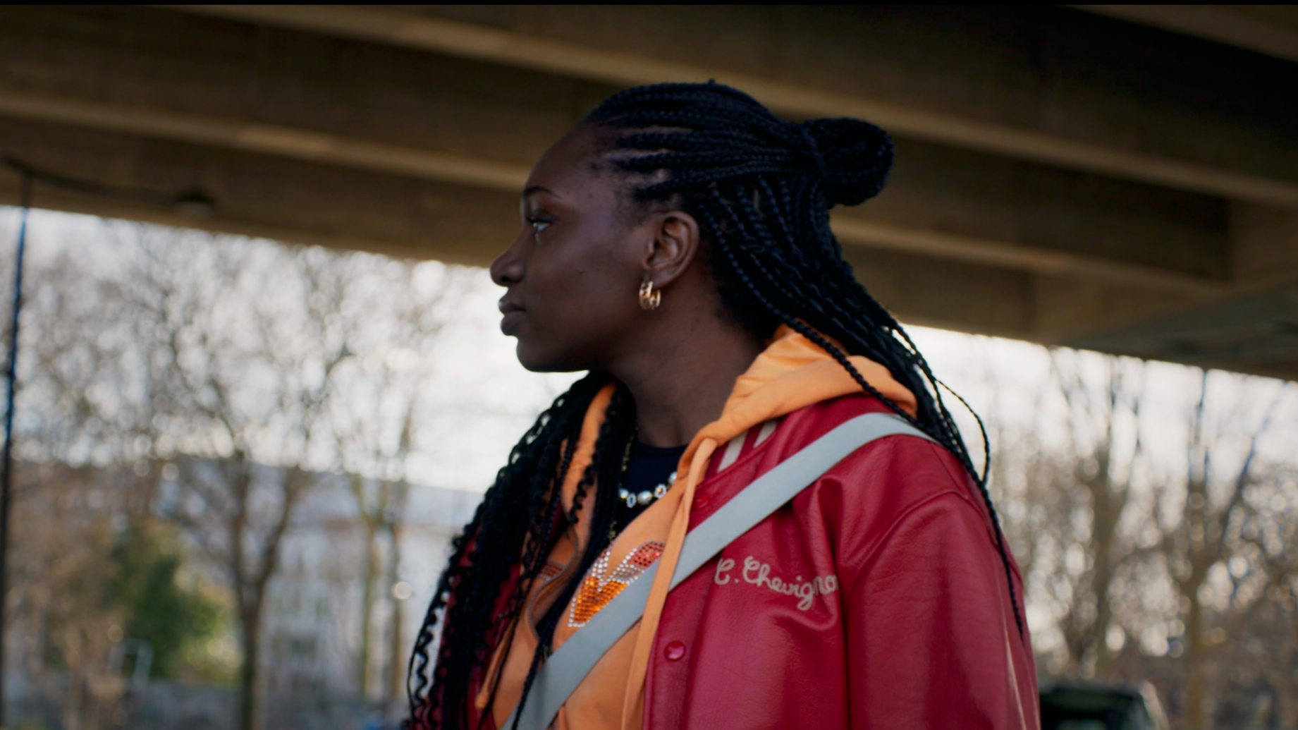 Une femme avec des cheveux tressés, portant un blouson rouge et une capuche orange, sous un pont en extérieur.