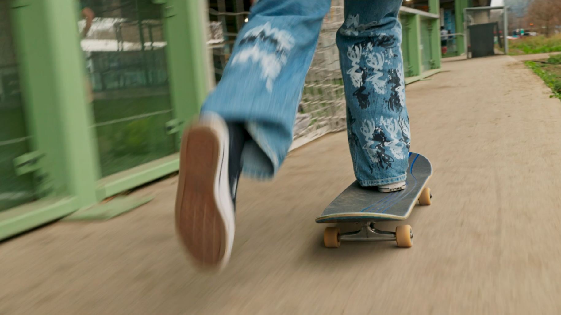 Une personne roulant en skateboard sur un chemin extérieur, portant un jeans à motifs et des chaussures de skate.