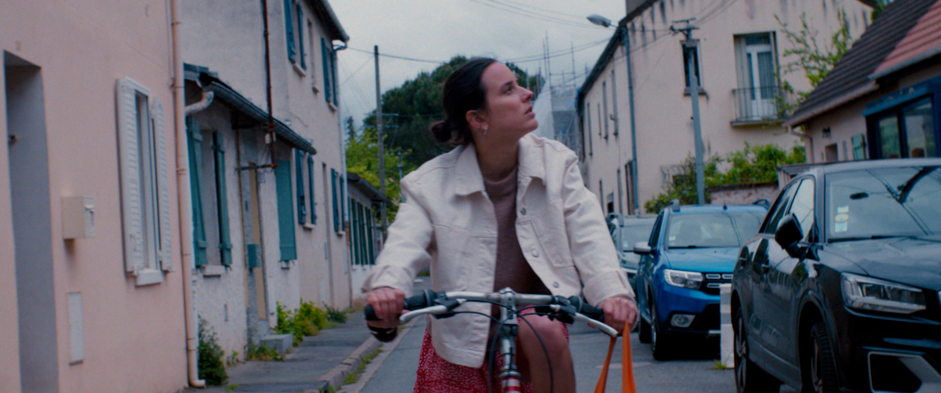 Une femme à vélo sur une rue résidentielle calme, entourée de maisons et de voitures, regardant vers le haut avec une expression contemplative.