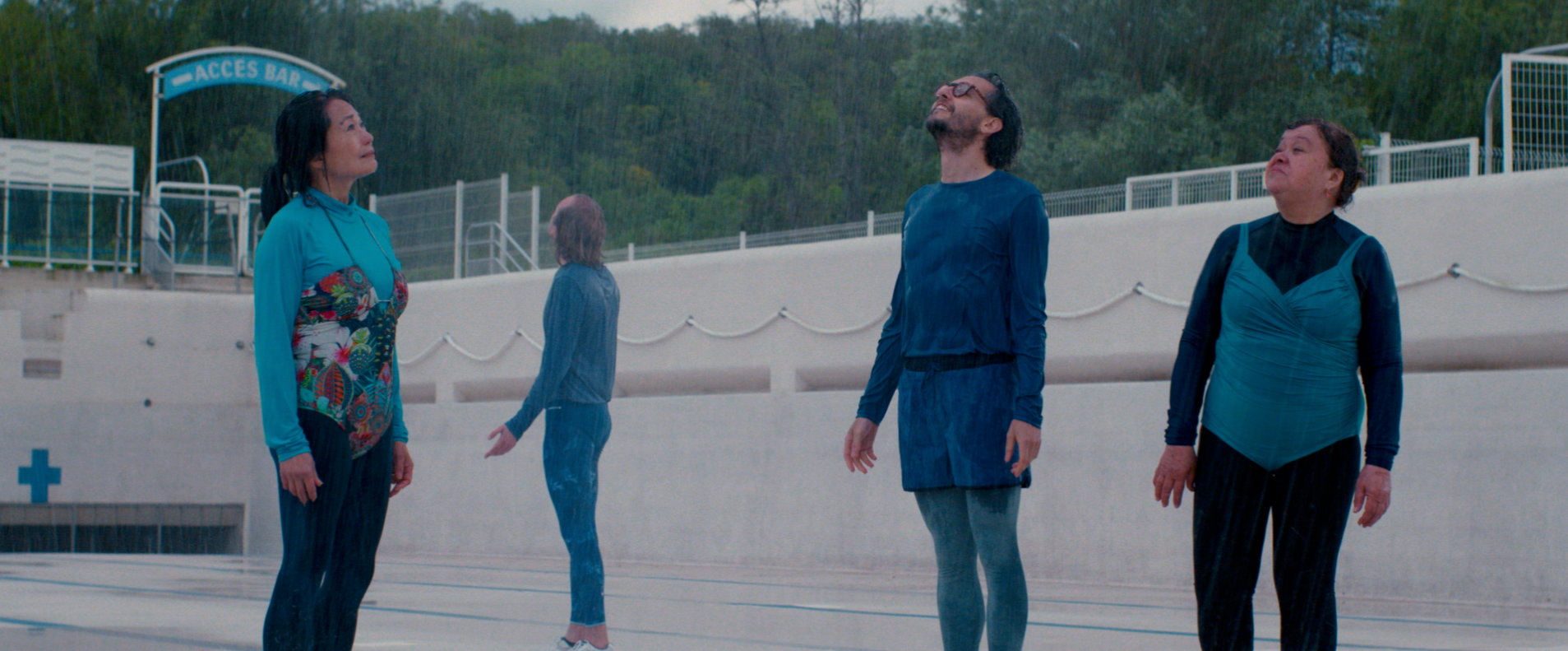 Quatre personnes sous la pluie, quelques-unes avec des vêtements colorés, regardent vers le haut dans une zone en dehors d'une piscine ou d'une installation sportive, avec un panneau indiquant 'Accès Bar' en arrière-plan.