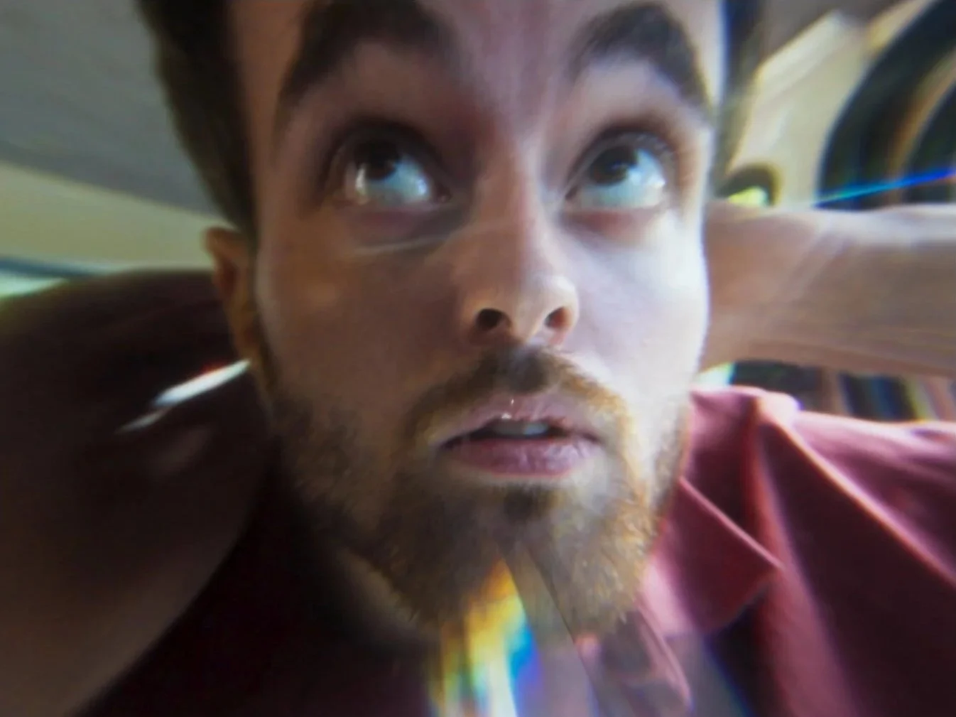 Un homme avec une barbe regarde vers le haut avec une expression surprise ou éveillée, portant une chemise rouge, dans un environnement intérieur.