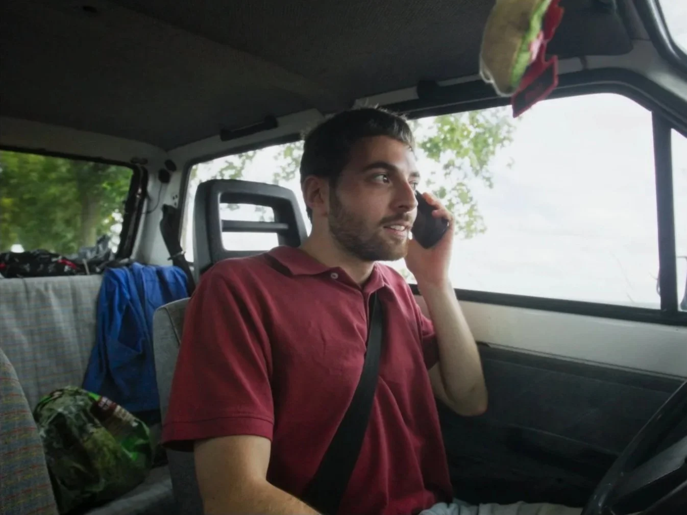 Un homme assis dans une camionnette, parlant au téléphone, avec un arbre visible par la fenêtre à l'arrière-plan.
