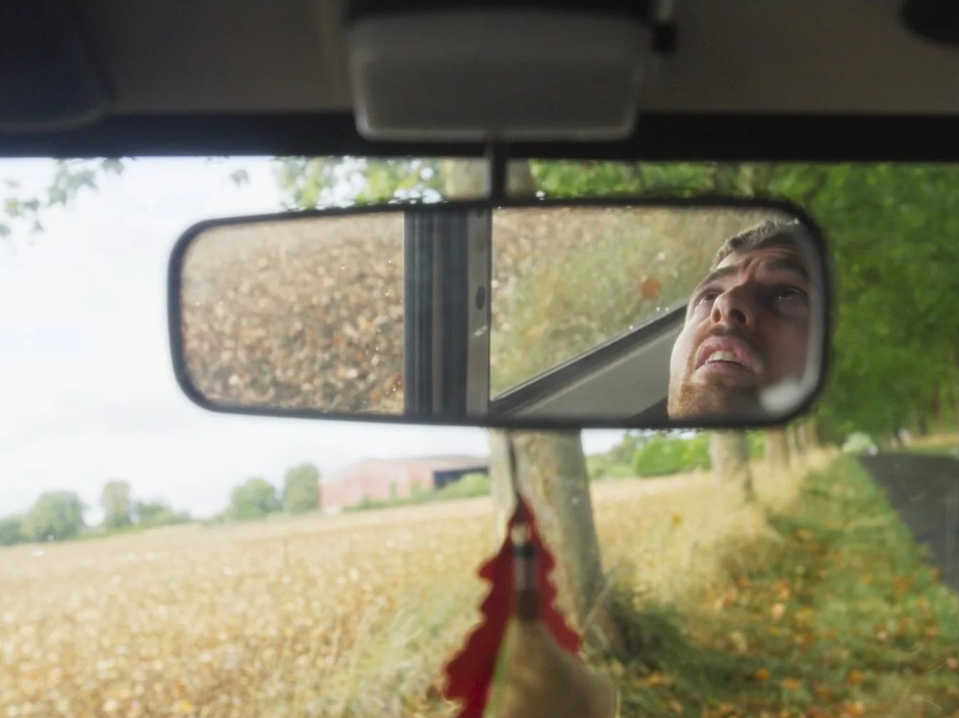 Reflet dans le miroir de rétroviseur d'un homme qui semble inquiet, avec un paysage bucolique en arrière-plan, comprenant des champs et des arbres.
