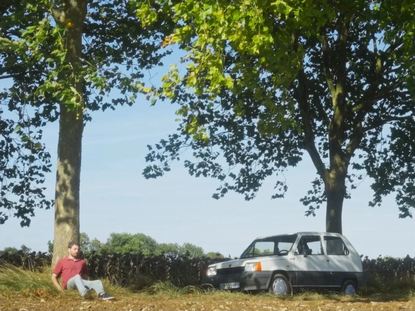Un homme assis sous un arbre à côté d'une voiture dans un champ en plein air.