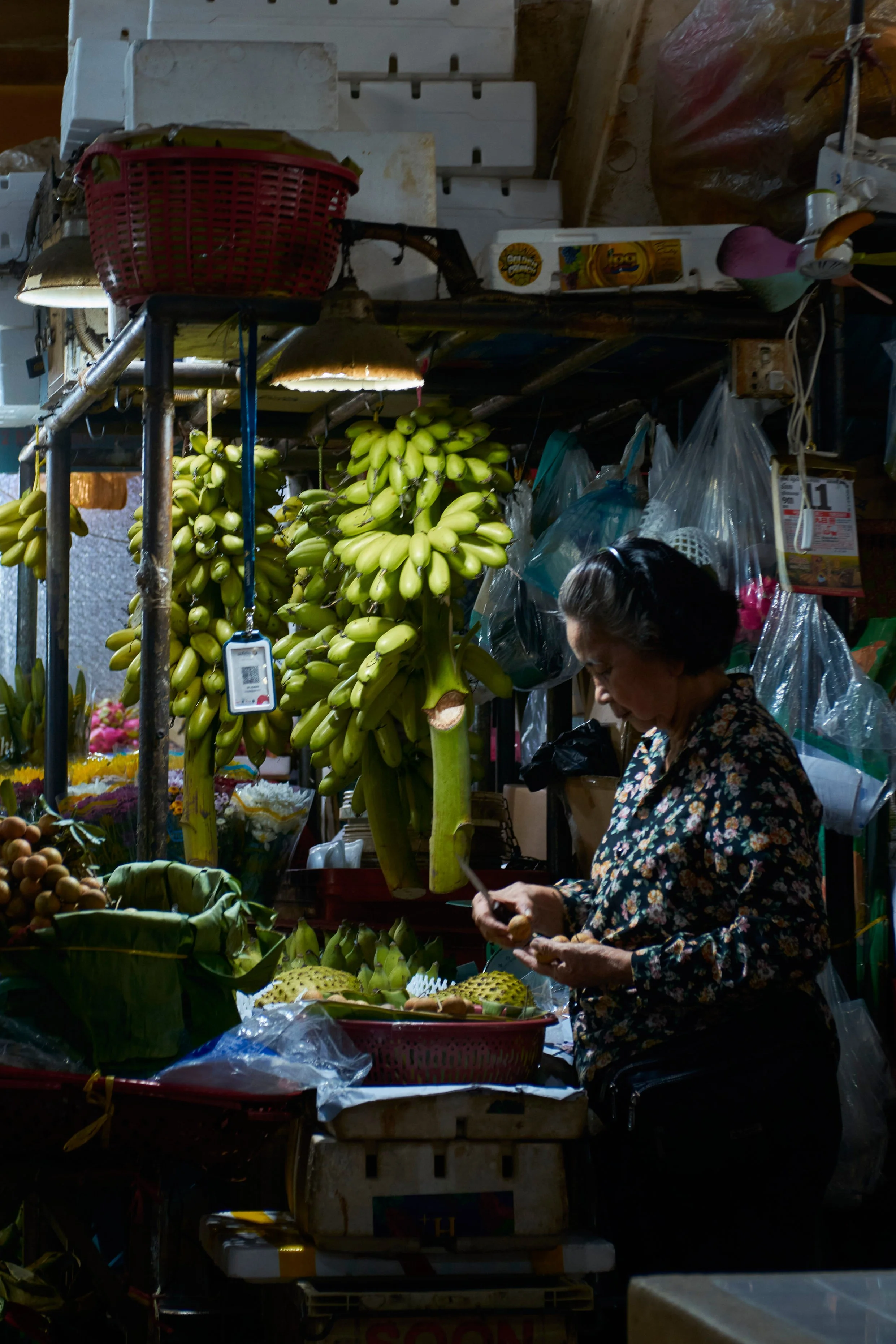 Une femme achète des bananes dans un marché couvert. Il y a de nombreux fruits et légumes, des sacs en plastique et des boîtes en carton, ainsi que des lampes éclairant l'étal.