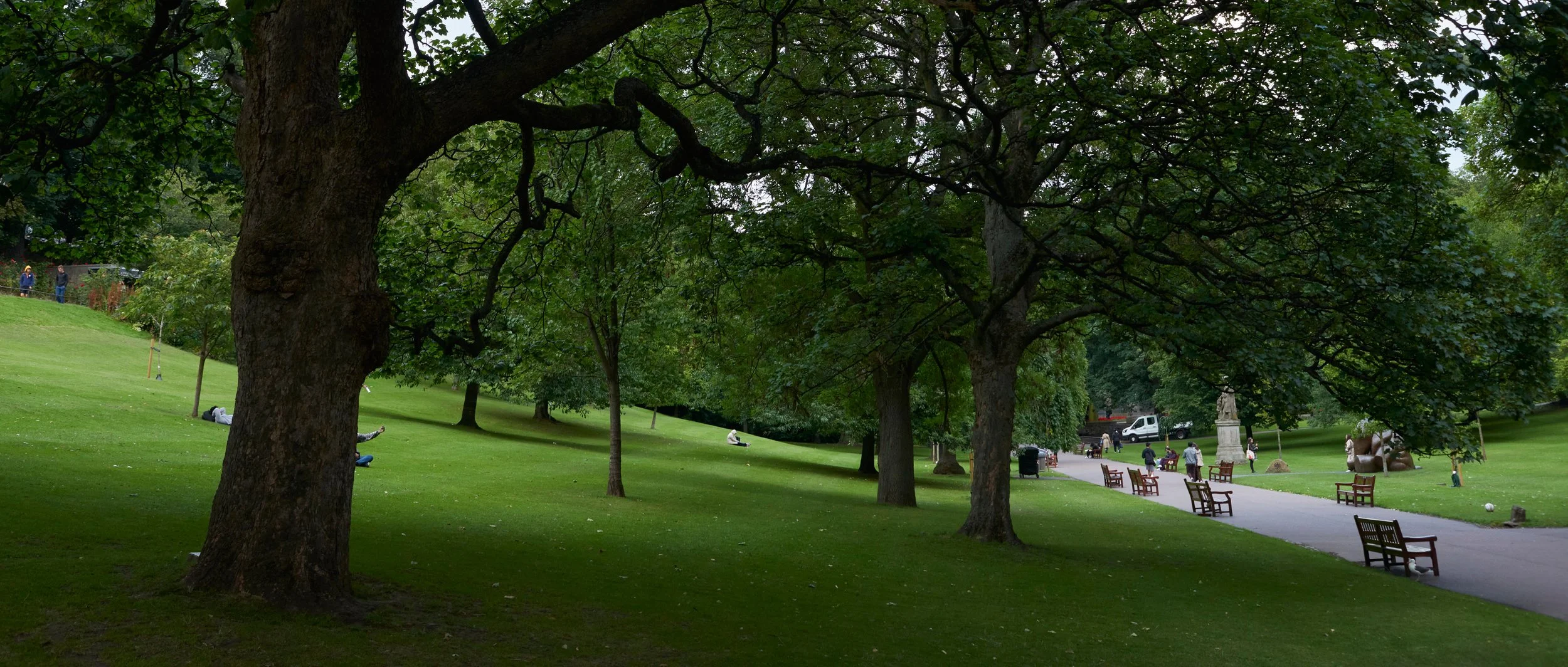 Un parc verdoyant avec des grands arbres, des chemins, des bancs, et quelques personnes se promenant ou se reposant.