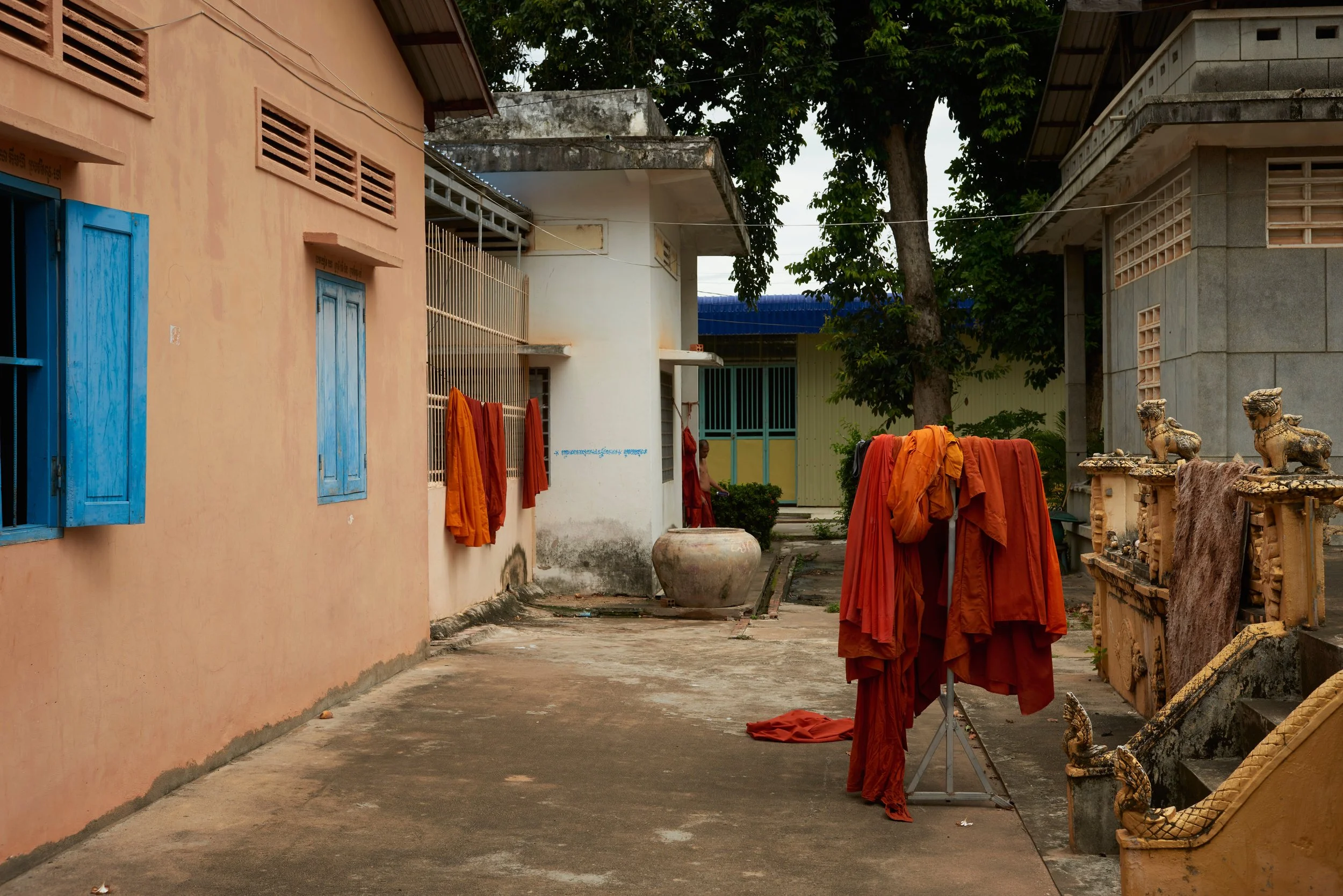 Cour d'une maison avec des robes de moine orange suspendues et des statues de lions en pierre à côté, murs en béton et arbres en arrière-plan.