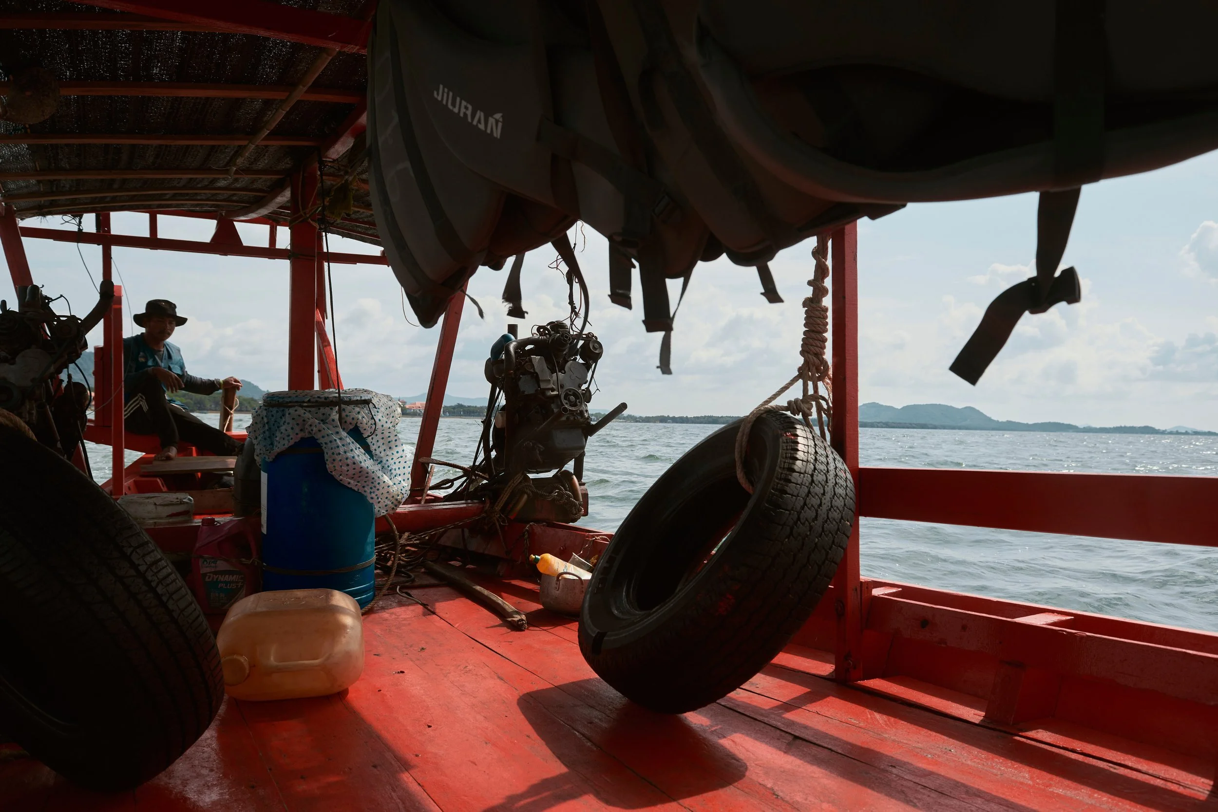 Vue depuis l'intérieur d'une jonque en bois rouge sur l'eau, avec des pneus de voiture comme rembourrage, un moteur, des bouées, des caisses, et des passagers regardant la mer avec le ciel en arrière-plan.
