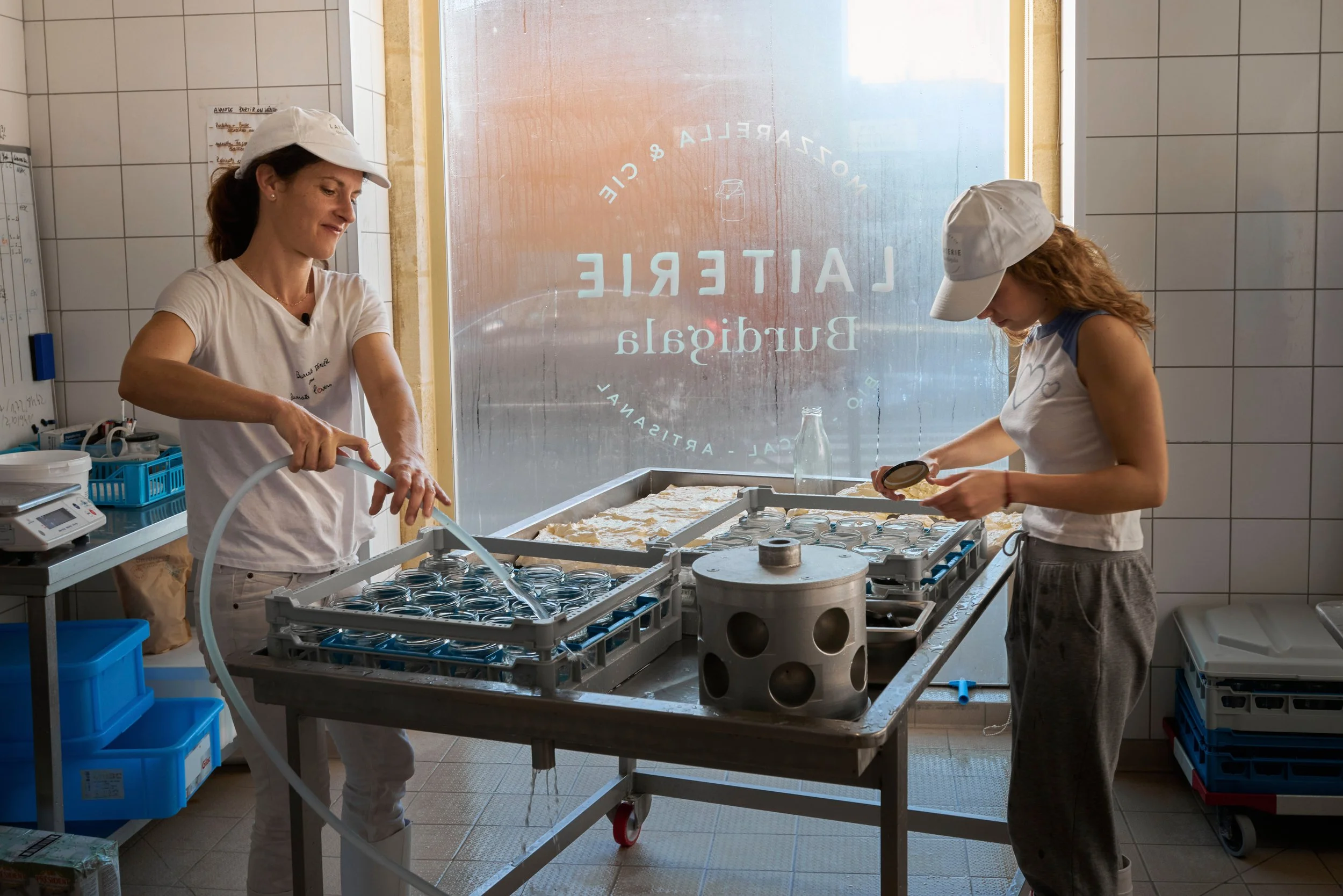 Deux femmes, l'une adulte et l'autre enfant, travaillent dans une cuisine avec des contenants en verre. Elles portent des casquettes blanches et semblent préparer ou stériliser des plats ou des bocaux.