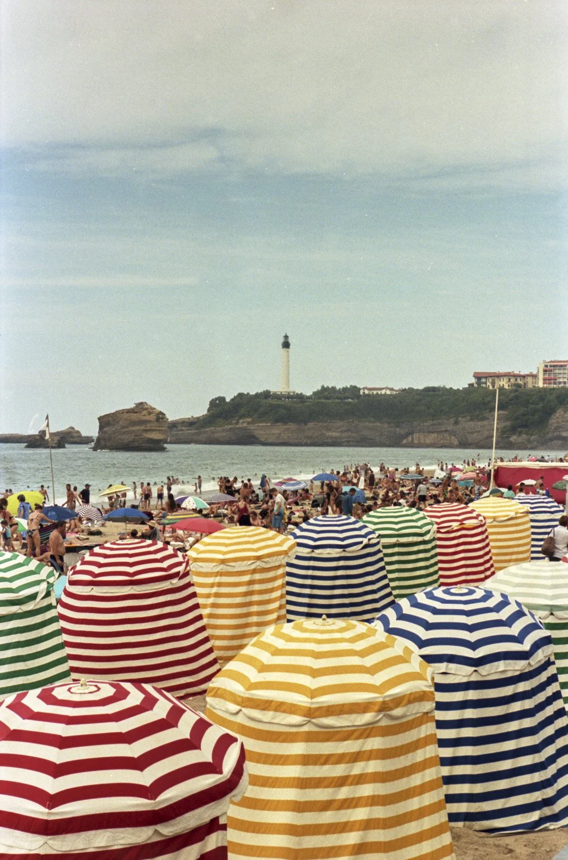 Plage bondée avec des parasols rayés colorés, mer calme, phare au loin, rochers et bâtiments en arrière-plan.