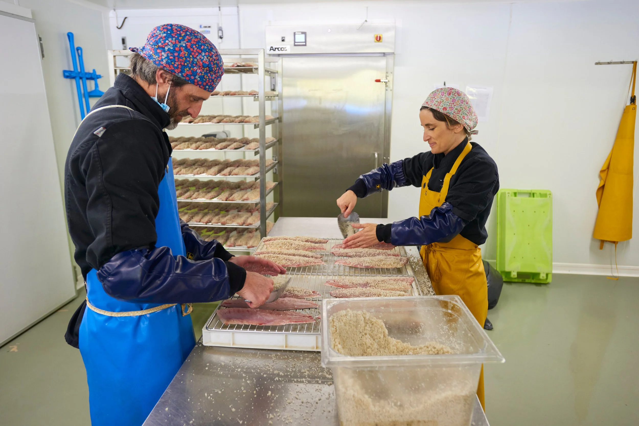 Deux personnes travaillent dans une cuisine ou un atelier, en train de préparer du poisson, avec des bacs contenant du riz ou de la chapelure. Elles portent des vêtements de protection, des gants et des bonnets pour maintenir l'hygiène.