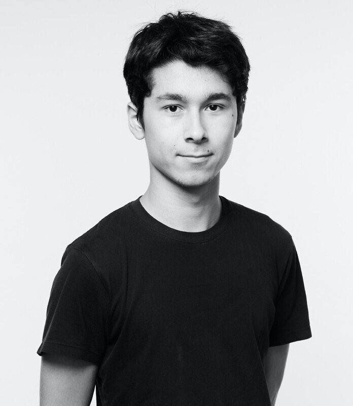 Portrait en noir et blanc d'un jeune homme avec des cheveux noirs et une expression neutre, portant un t-shirt noir, photo prise devant un fond blanc.