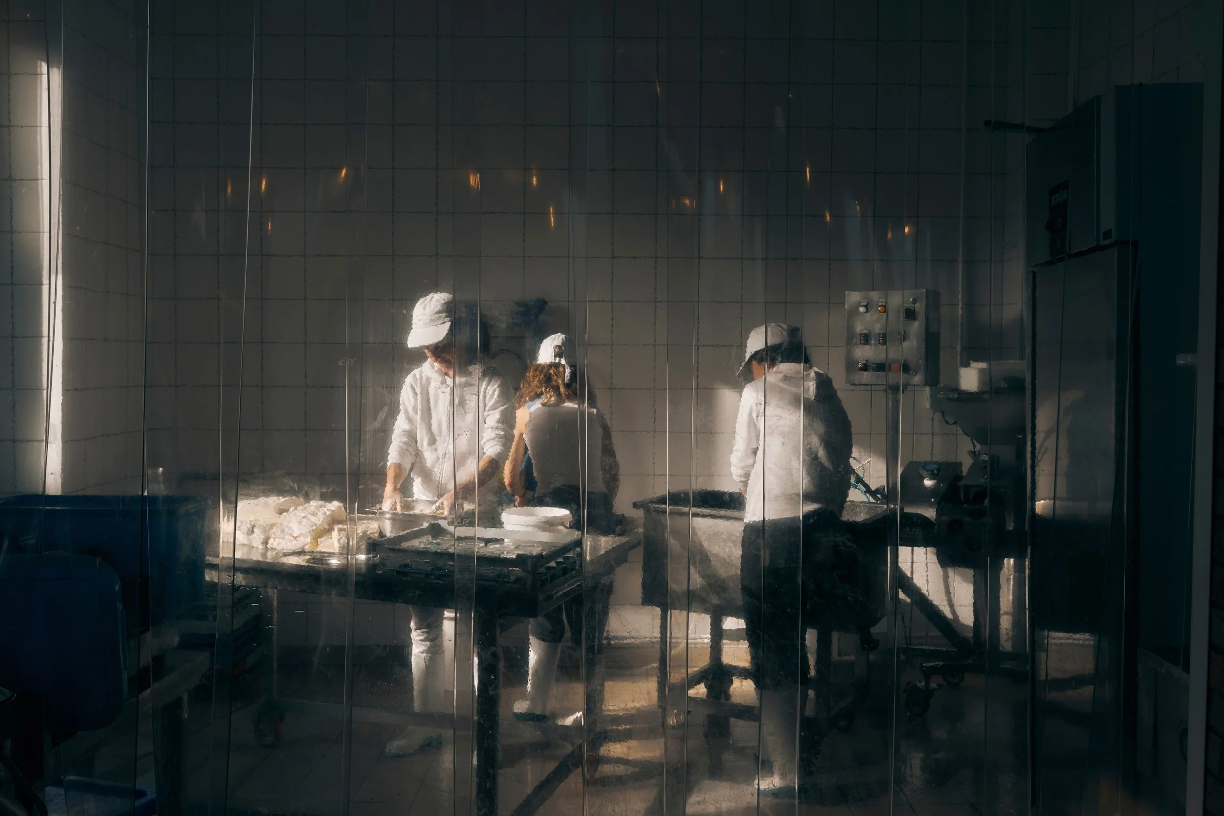 Trois personnes habillées en blanc, portant des chapeaux, travaillent derrière une vitre à nettoyer ou retirer de la matière blanche, probablement de la pâte ou de la mousse, dans un environnement de cuisine ou de laboratoire.