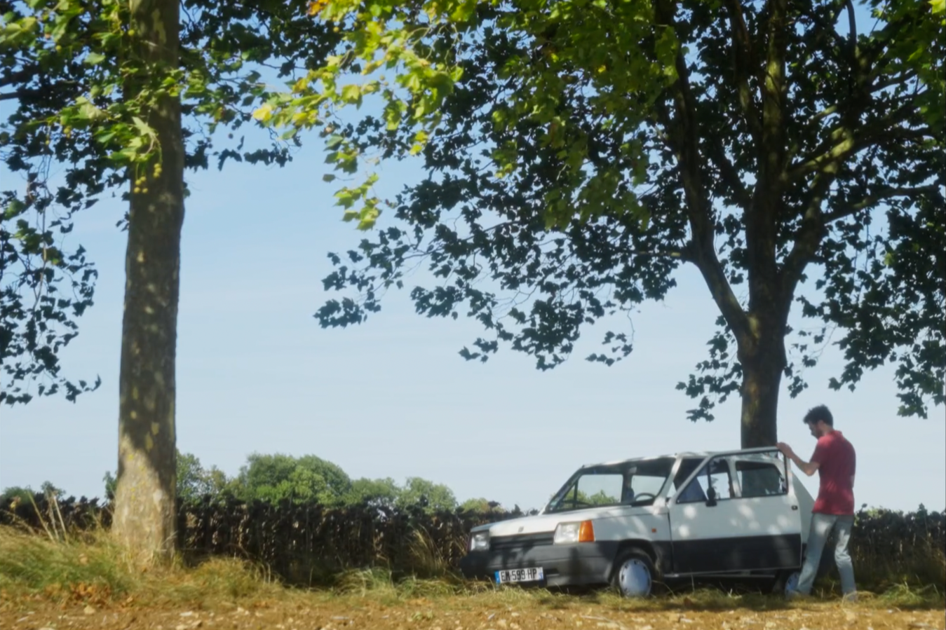 Un homme à côté d'une voiture blanche arrêtée sous un arbre dans un paysage rural ensoleillé.