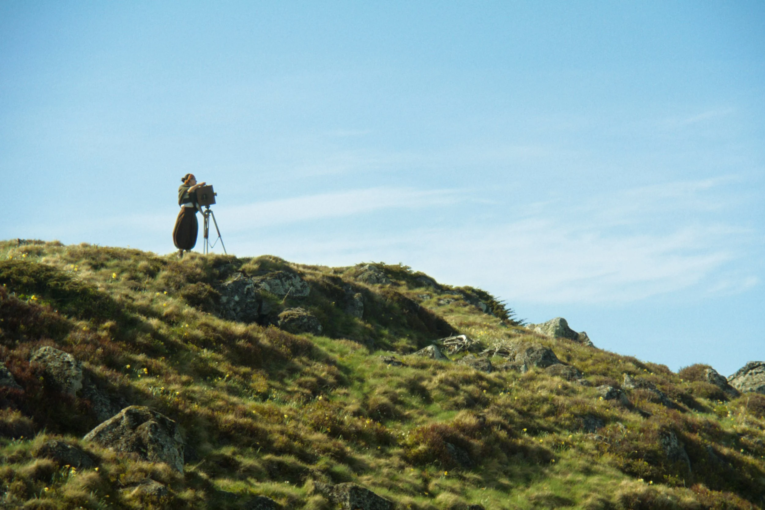 Une personne caminant avec un chapeau et un sac, utilisant un télescope ou un appareil photo sur un trépied sur une colline verdoyante sous un ciel bleu clair.