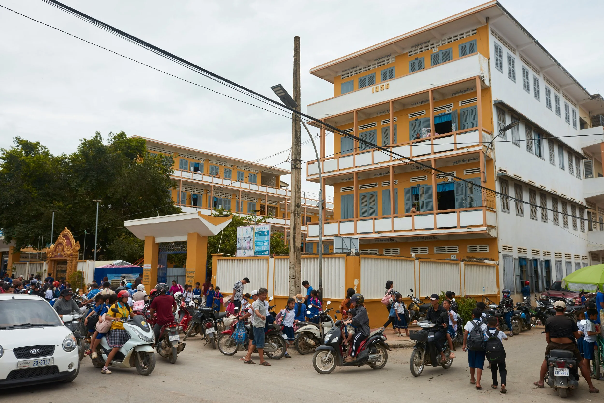 Une foule de personnes, dont beaucoup d'enfants en uniforme scolaire, se rassemble devant une école en architecture moderne, aux murs orange et bleus, avec plusieurs étages et fenêtres à volets. Beaucoup de motos et quelques voitures sont stationnées