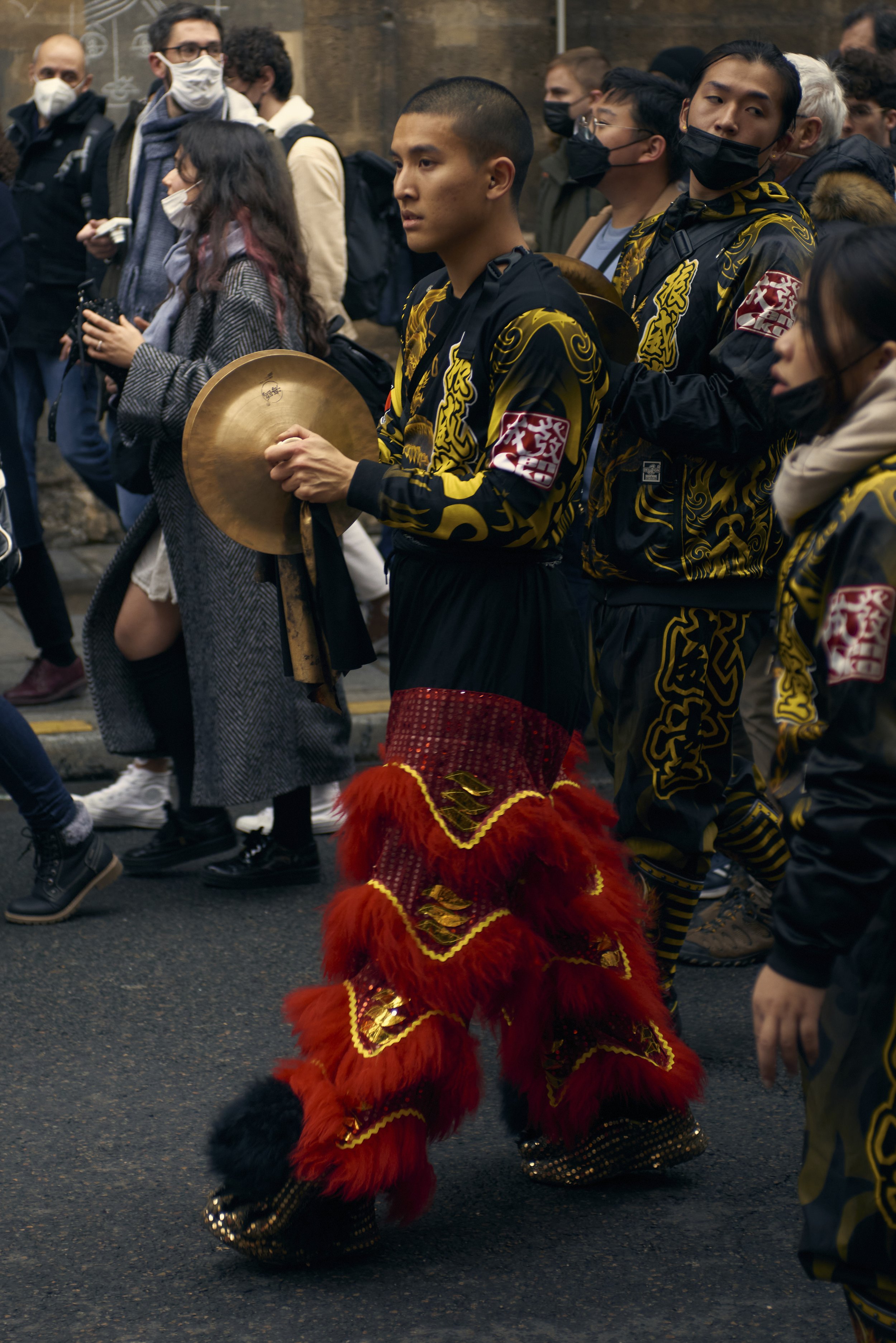 Danseurs traditionnels asiatiques vêtu de costumes noirs et or, portant des masques noirs, en participation à une parade ou festival dans une rue urbaine, avec une foule de spectateurs