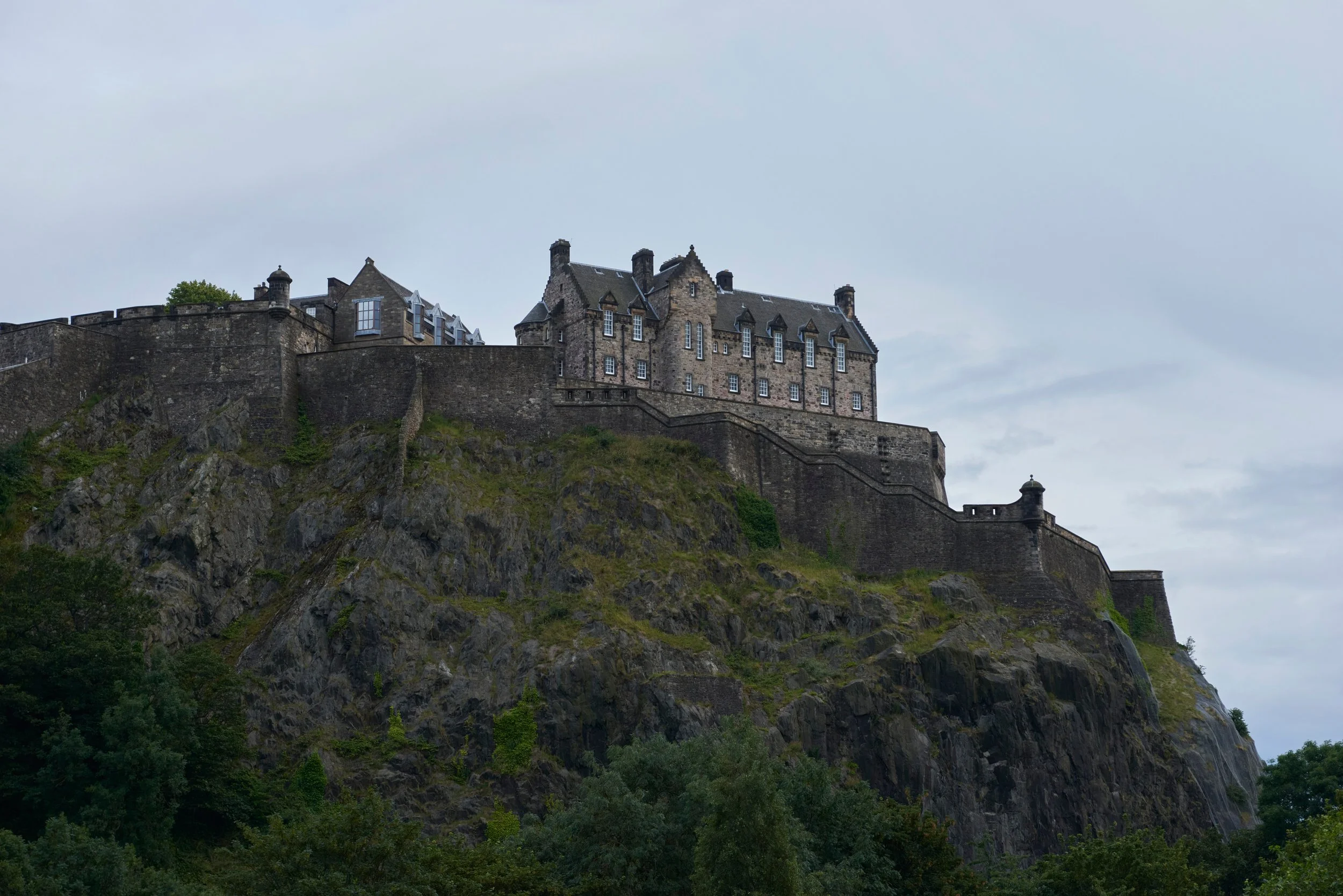 Un château ancien en pierre situé au sommet d'une colline rocheuse, entouré de végétation verdoyante, sous un ciel nuageux.