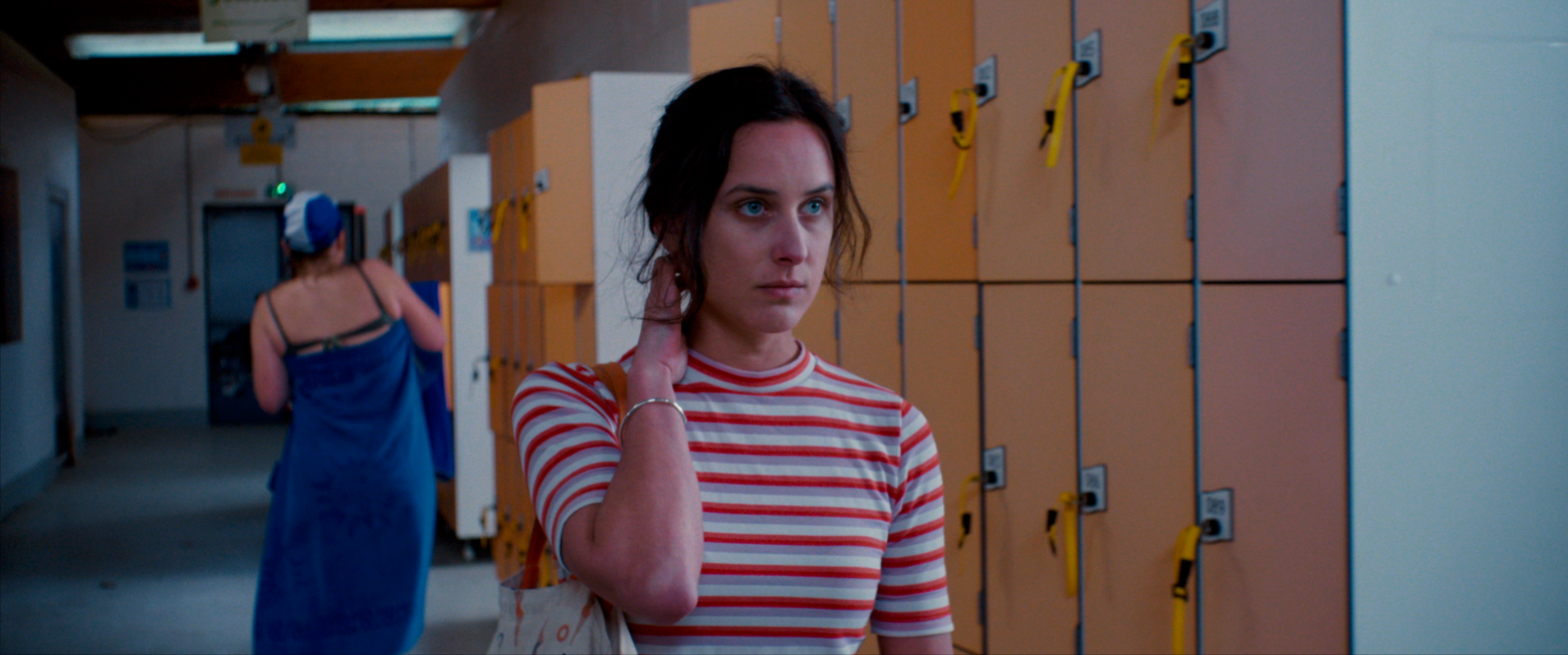 Jeune femme dans une salle de vestiaires, portant un t-shirt à rayures rouges et blanches, semblant préoccupée, avec des casiers orange en arrière-plan.