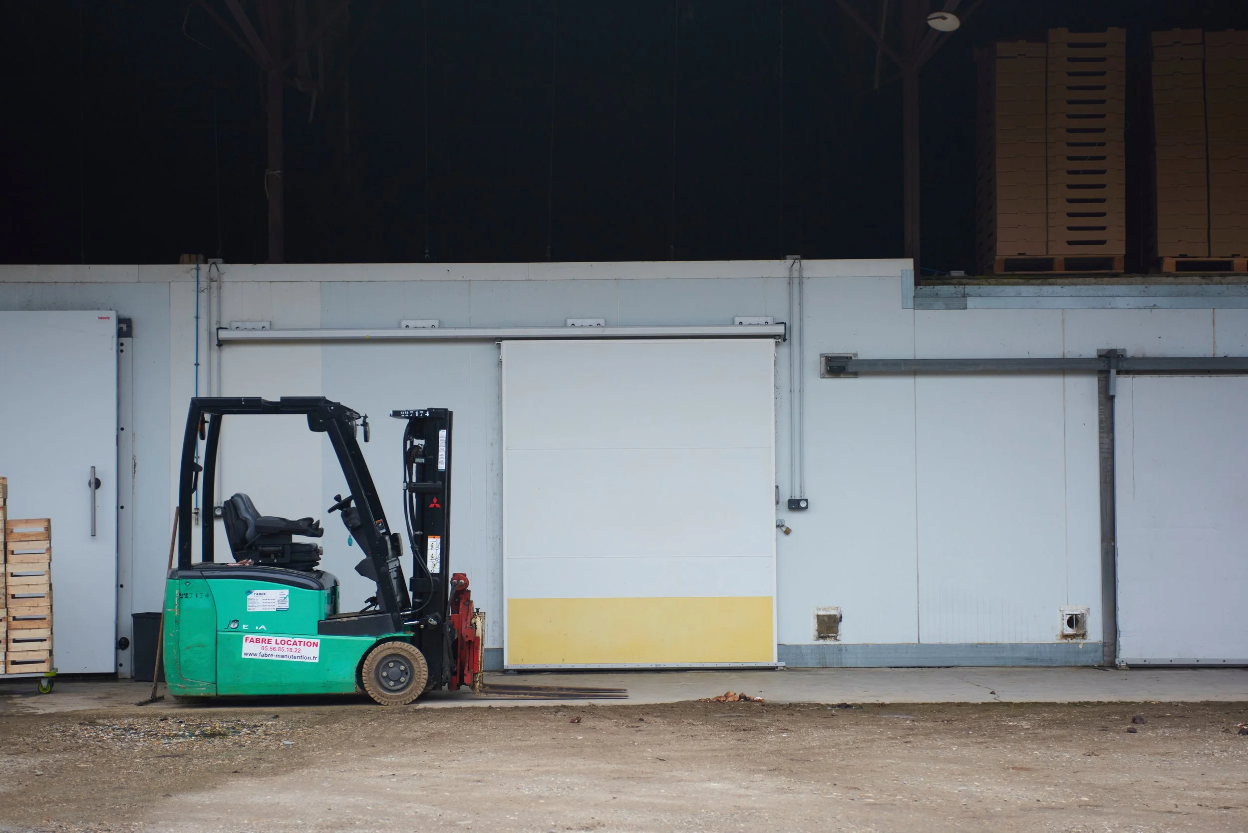 Un chariot élévateur vert garé devant un bâtiment blanc industriel avec une grande porte métallique et des palettes en bois empilées. La scène se déroule à l'extérieur, sur un sol en terre ou gravier.