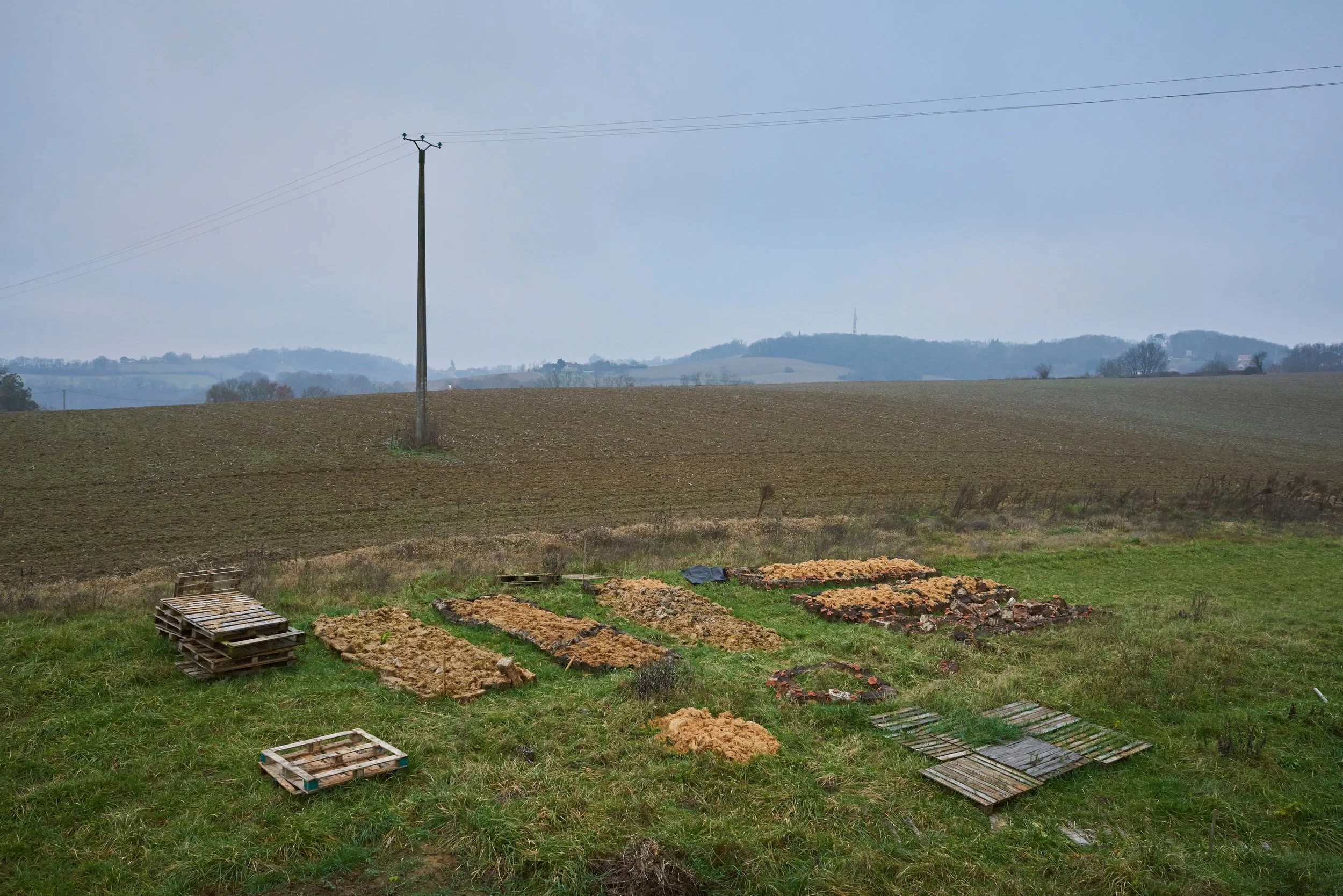 Champs agricoles avec palettes en bois, piles de terre, et zones cultivées, sous un ciel nuageux, avec des collines en arrière-plan.