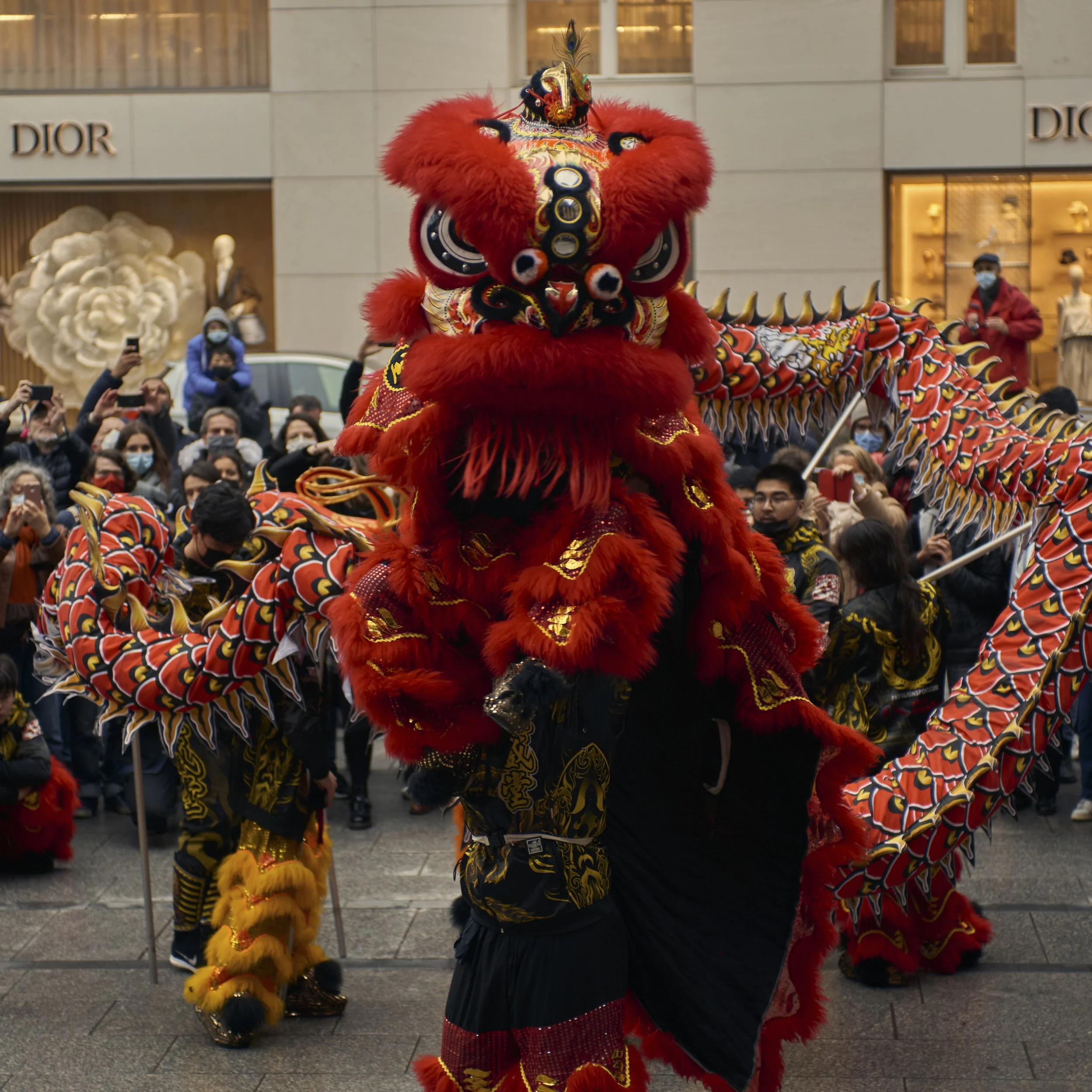 Un festival avec une danse traditionnelle chinoise du lion rouge dans une rue urbaine, avec des spectateurs portant des masques et des magasins de luxe en arrière-plan.
