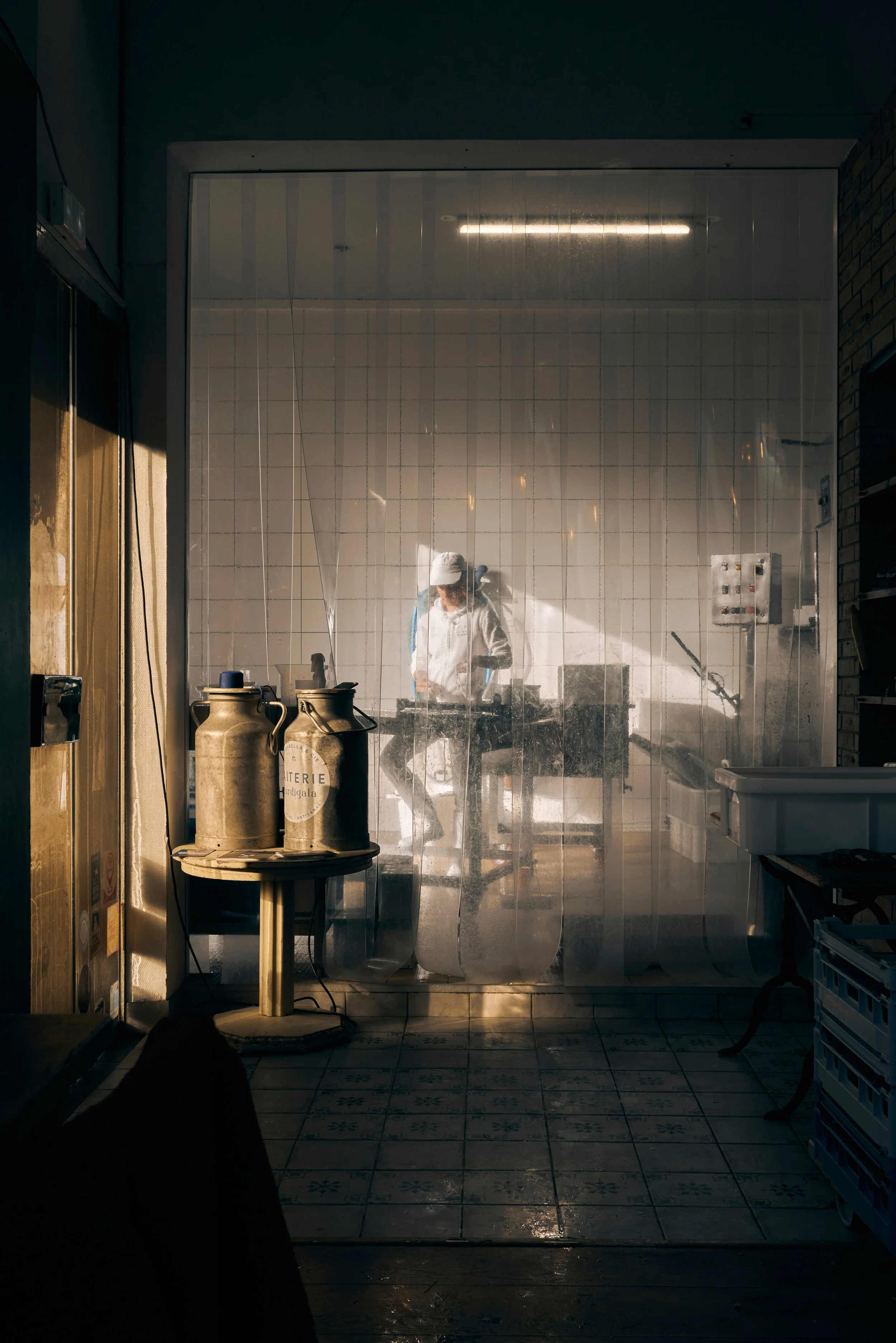 Une jeune femme portant une blouse et un bonnet blanc est assise dans une cuisine derrière une vitre en plastique. La cuisine comporte un évier, des bonbonnes, des ustensiles et un panneau électrique. La pièce est faiblement éclairée, et la lumière n
