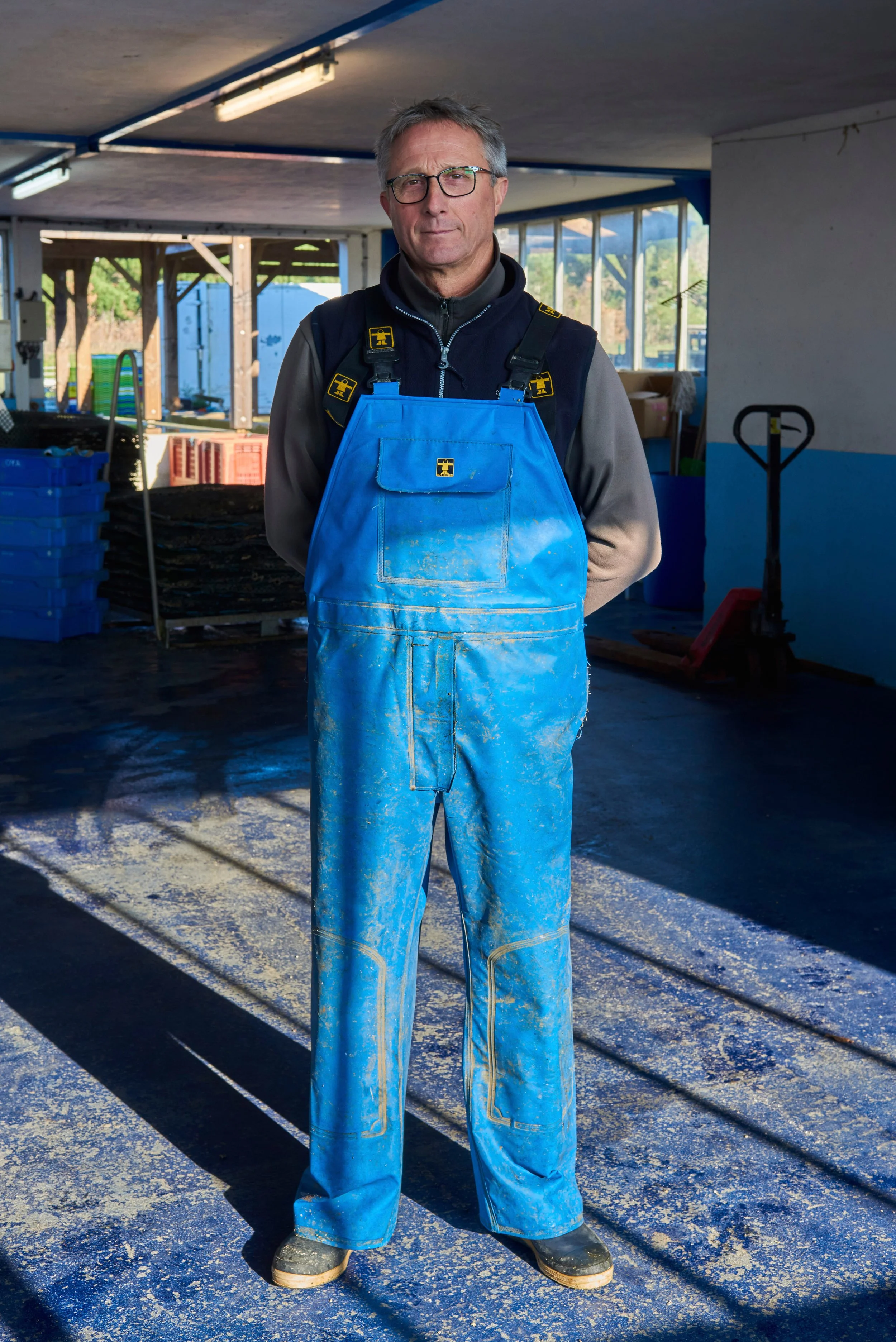 Un homme porte une combinaison de travail bleue avec des gants et des lunettes de sécurité, se tenant dans un espace intérieur avec des fenêtres, du matériel de construction et un sol poussiéreux.