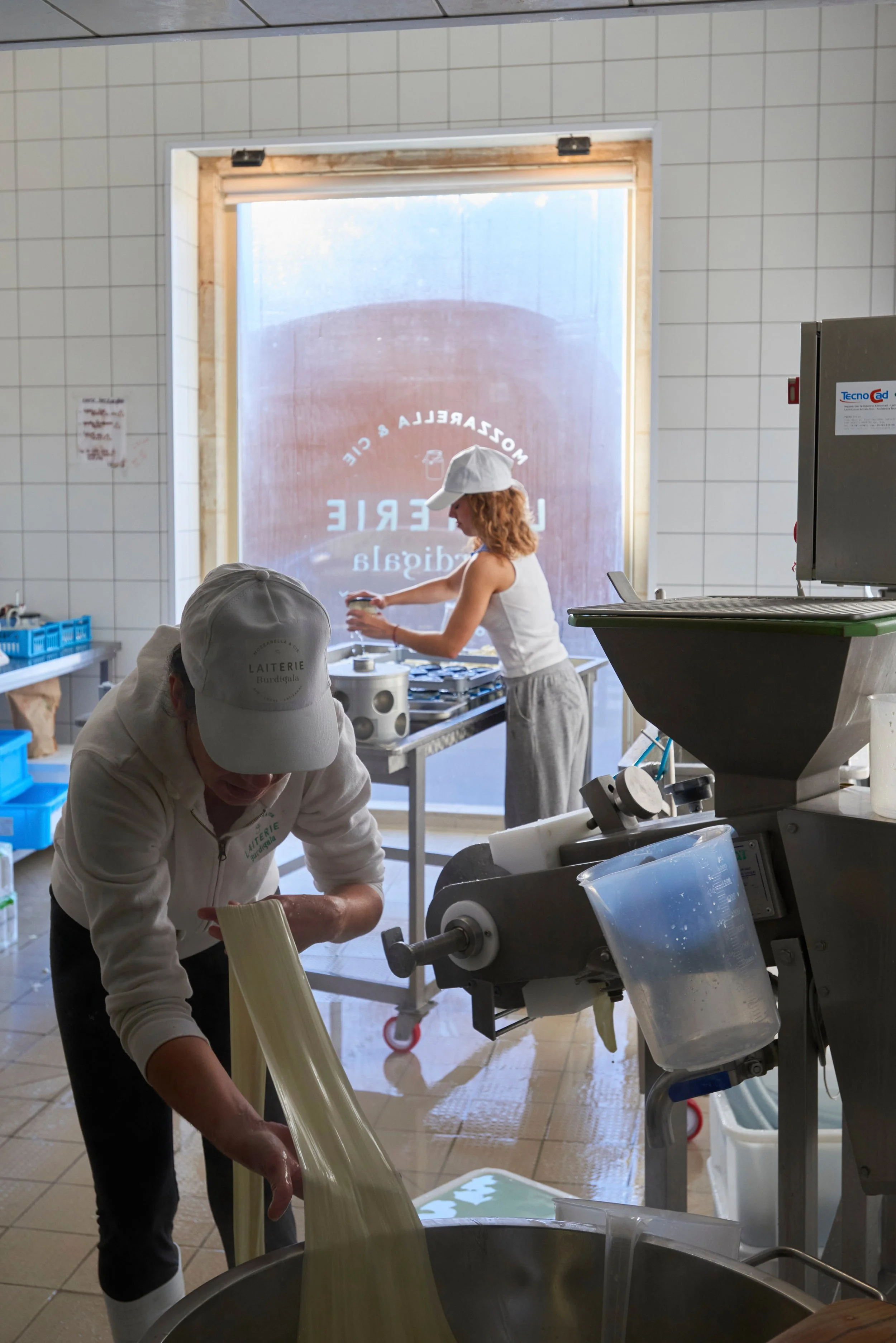 Deux personnes, vêtues de vêtements blancs et portant des casquettes blanches, travaillent dans une cuisine ou une usine de fabrication de fromage ou de produits laitiers. Elles manipulent de longues bandes de fromage ou de pâte qui pendent dans une 