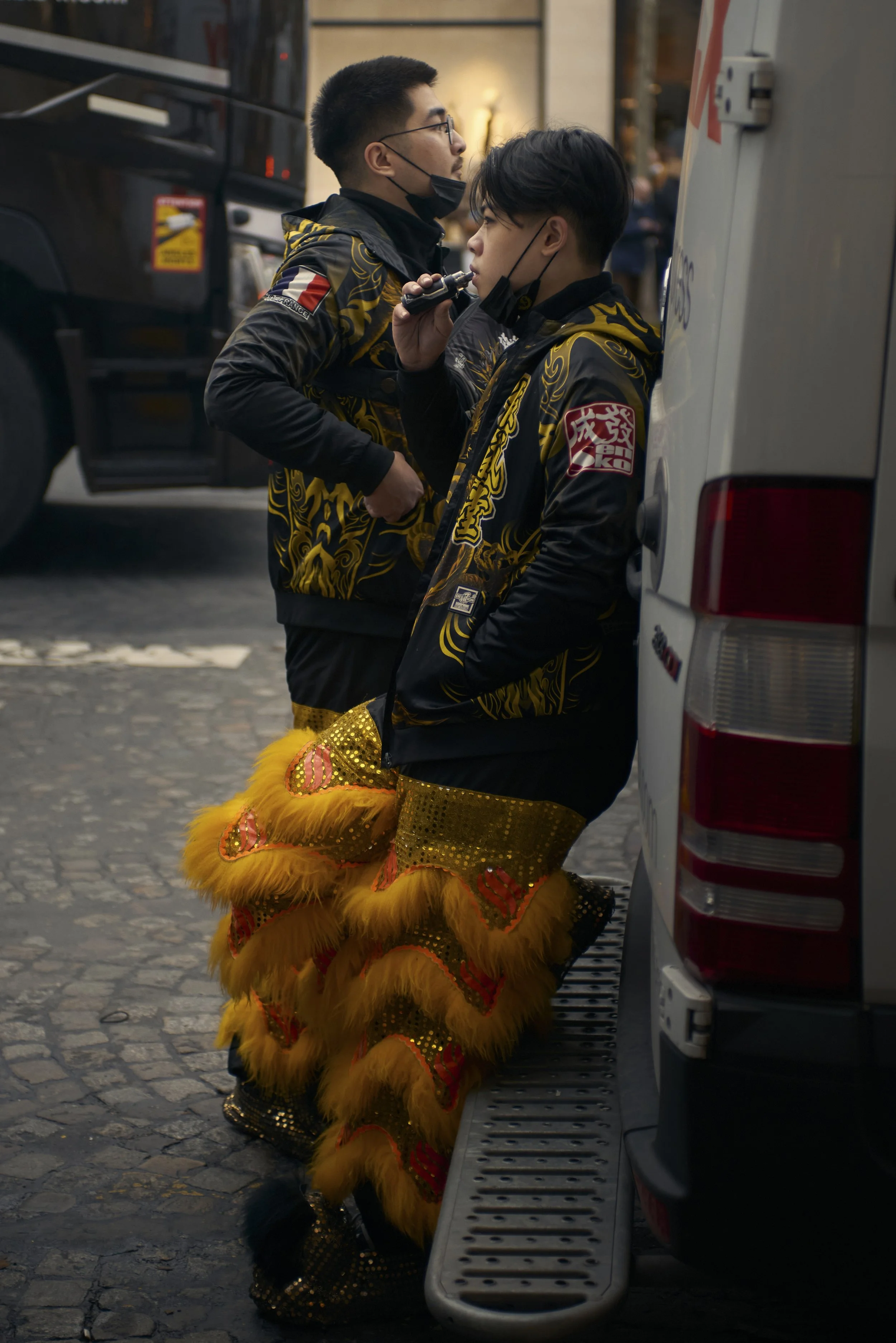 Deux jeunes hommes portant des costumes de lion traditionnels chinois sont debout contre un véhicule blanc, discutant. L'un d'eux tient une radio, ils portent tous deux des masques faciaux noirs abaissés sous le menton. La scène se déroule en milieu 