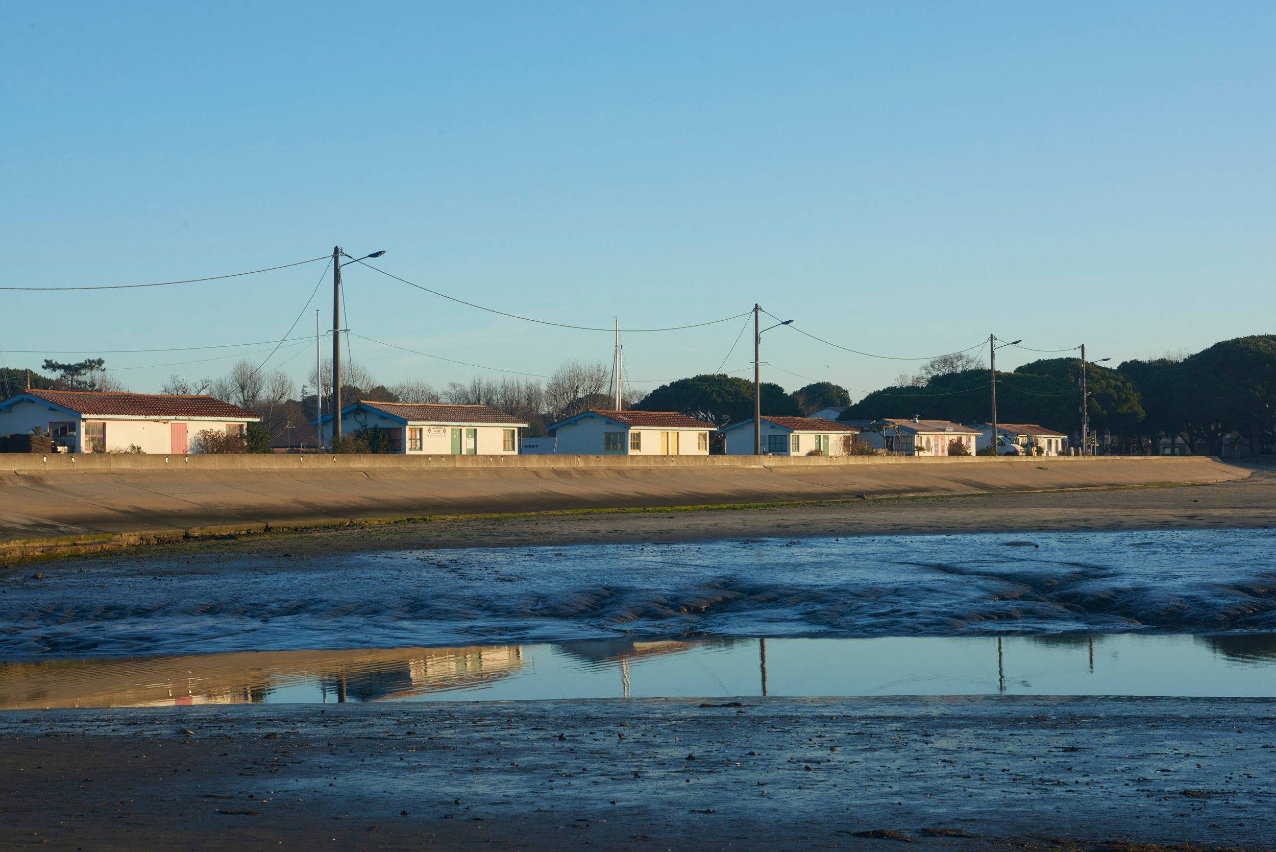 Petites maisons colorées avec des toits en tuiles rouges, alignées le long d'une voie en bordure d'eau, avec des pylônes électriques et un ciel dégagé.