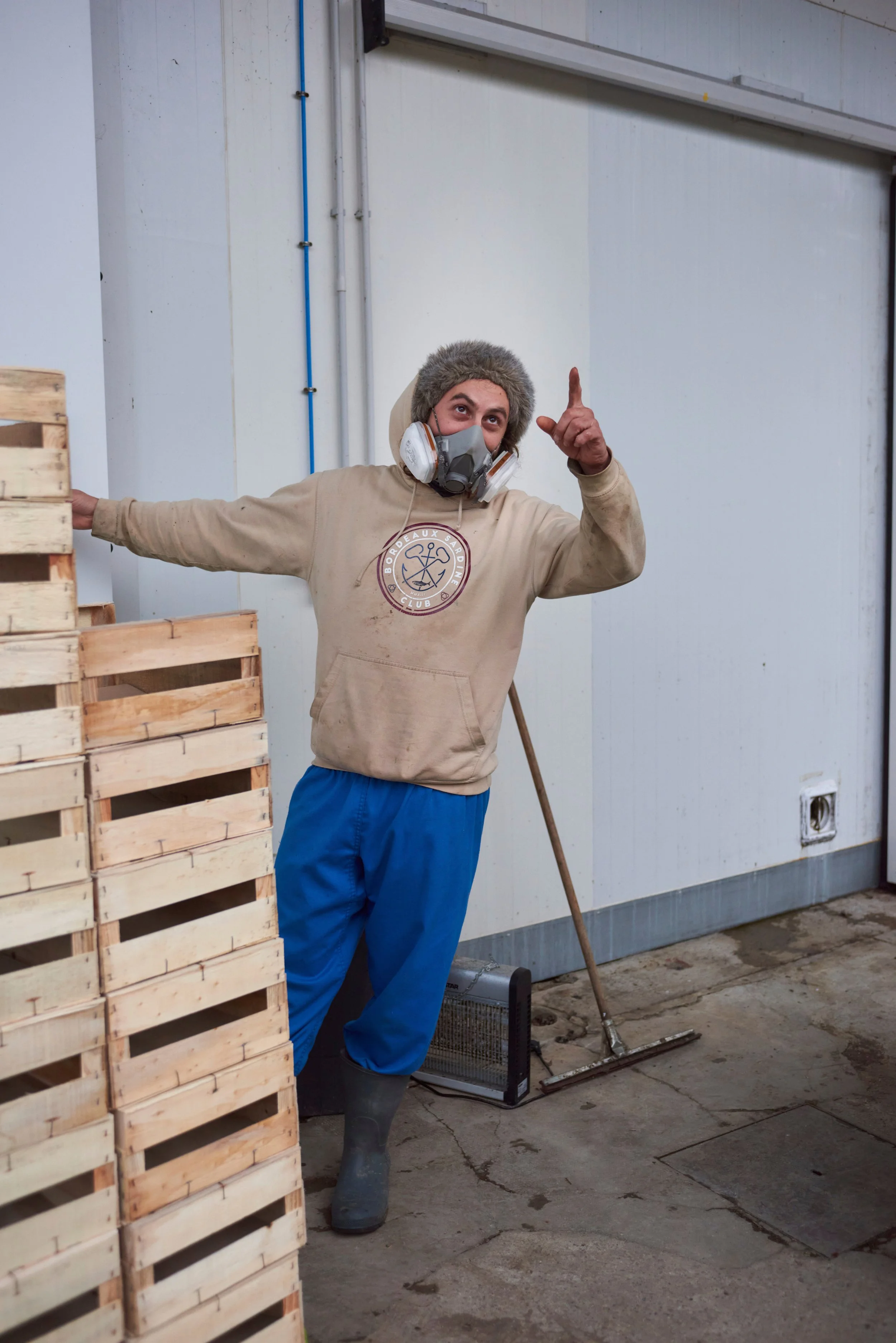 Un homme portant un sweat-shirt beige avec un logo et des bottes en caoutchouc, portant un masque respiratoire, se tient à côté d'une pile de palettes en bois, levant le doigt en l'air dans un entrepôt ou une usine.