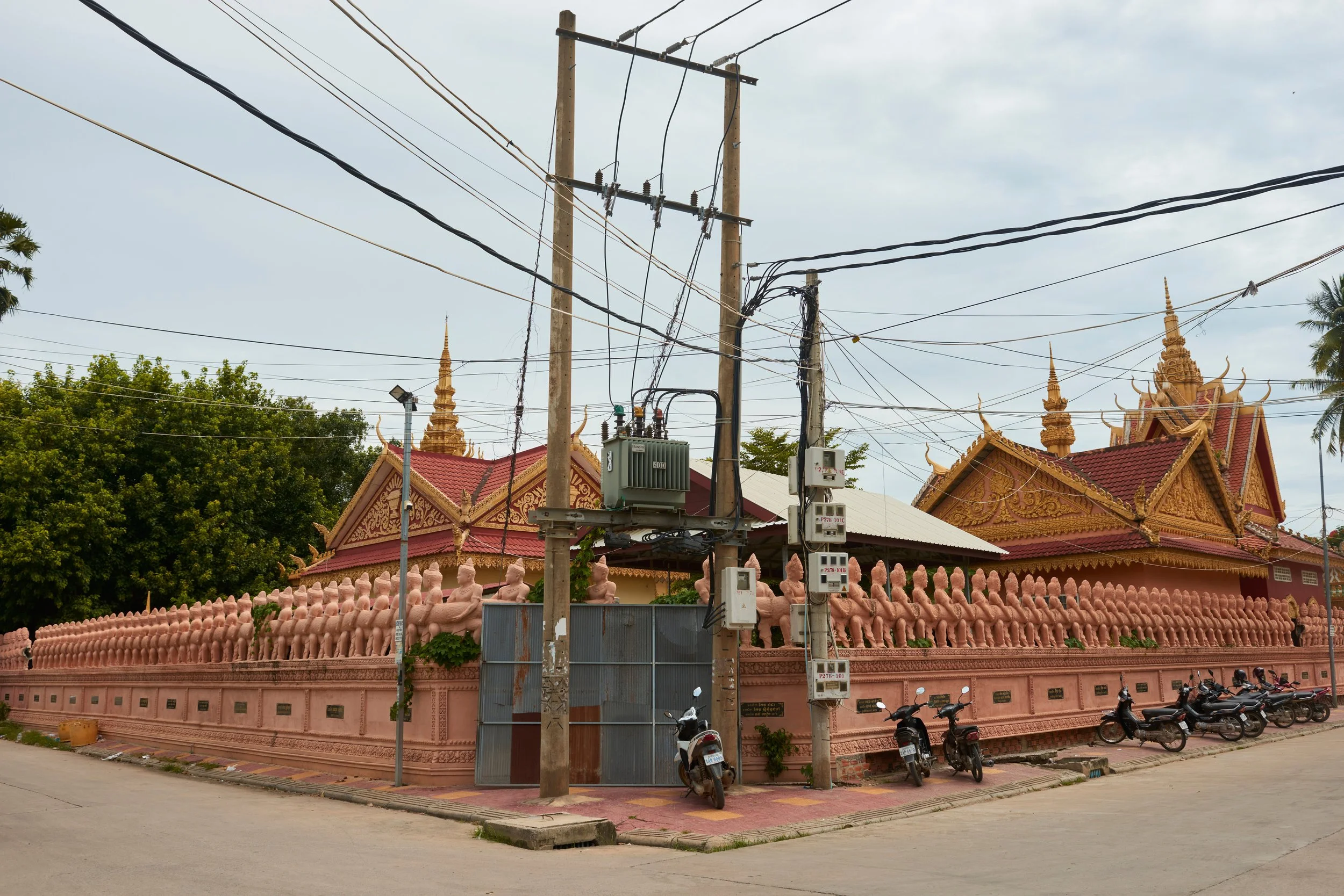 Temple bouddhiste avec une clôture rose, des statues de Bouddha, des arbres verts, un poteau électrique, une ligne électrique suspendue, motos garées sur le trottoir, un ciel nuageux.